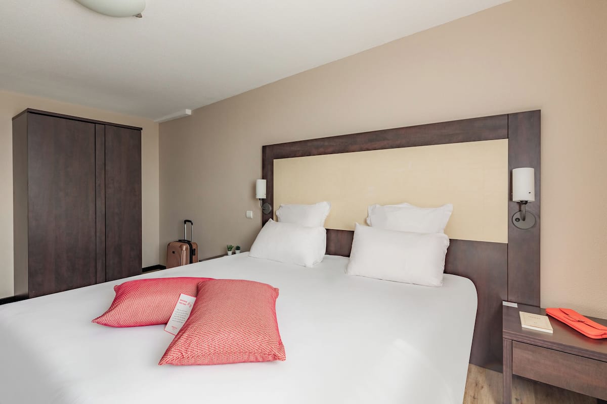 A neatly made bed with white and red pillows, a wooden headboard, and a nightstand with a lamp and a book.