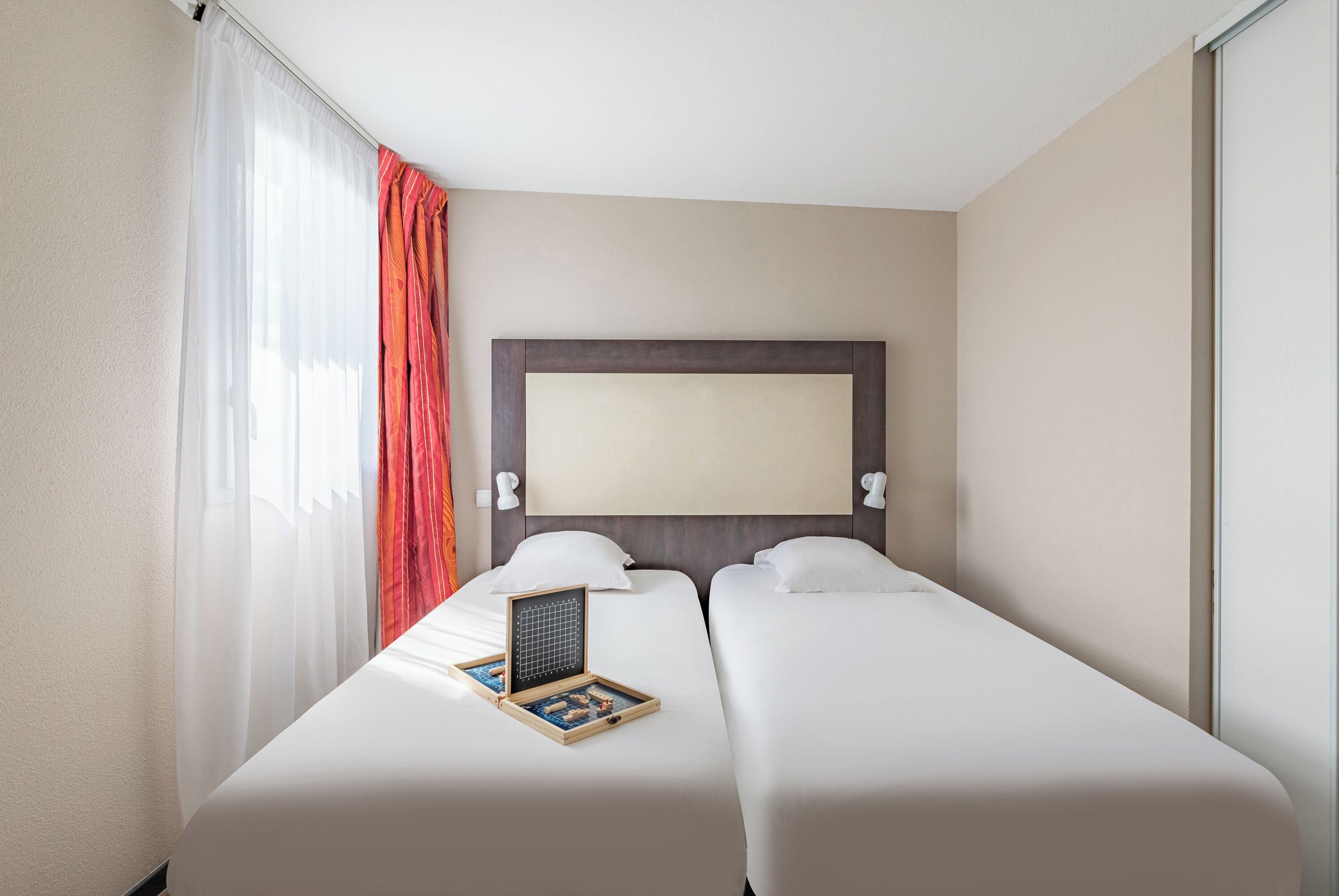 Habitación de hotel con dos camas, un ventanal con cortinas translúcidas, cabecera y una mesita con un juego de mesa.