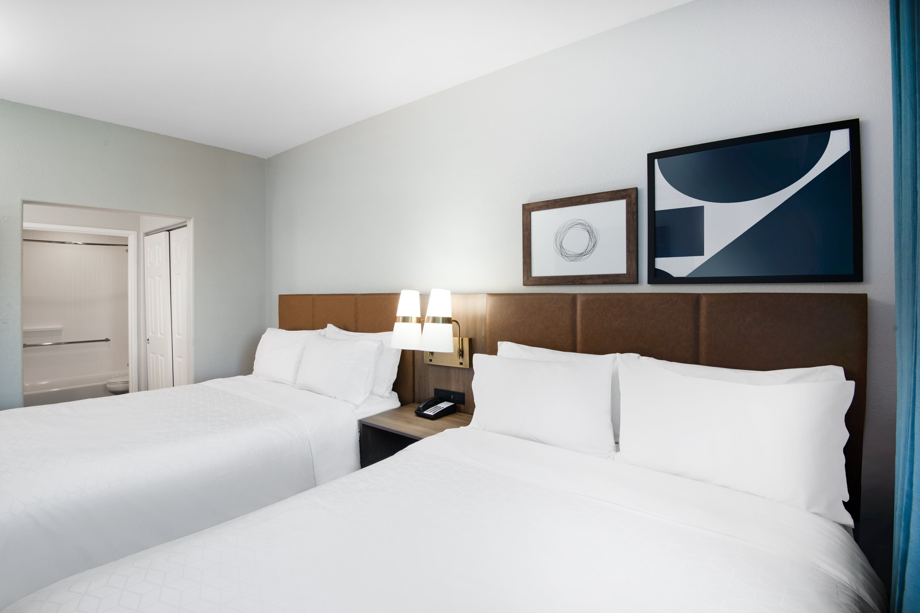 Une chambre d’hôtel avec deux lits, une table de chevet avec une lampe, un tableau encadré et une porte donnant sur une salle de bain.