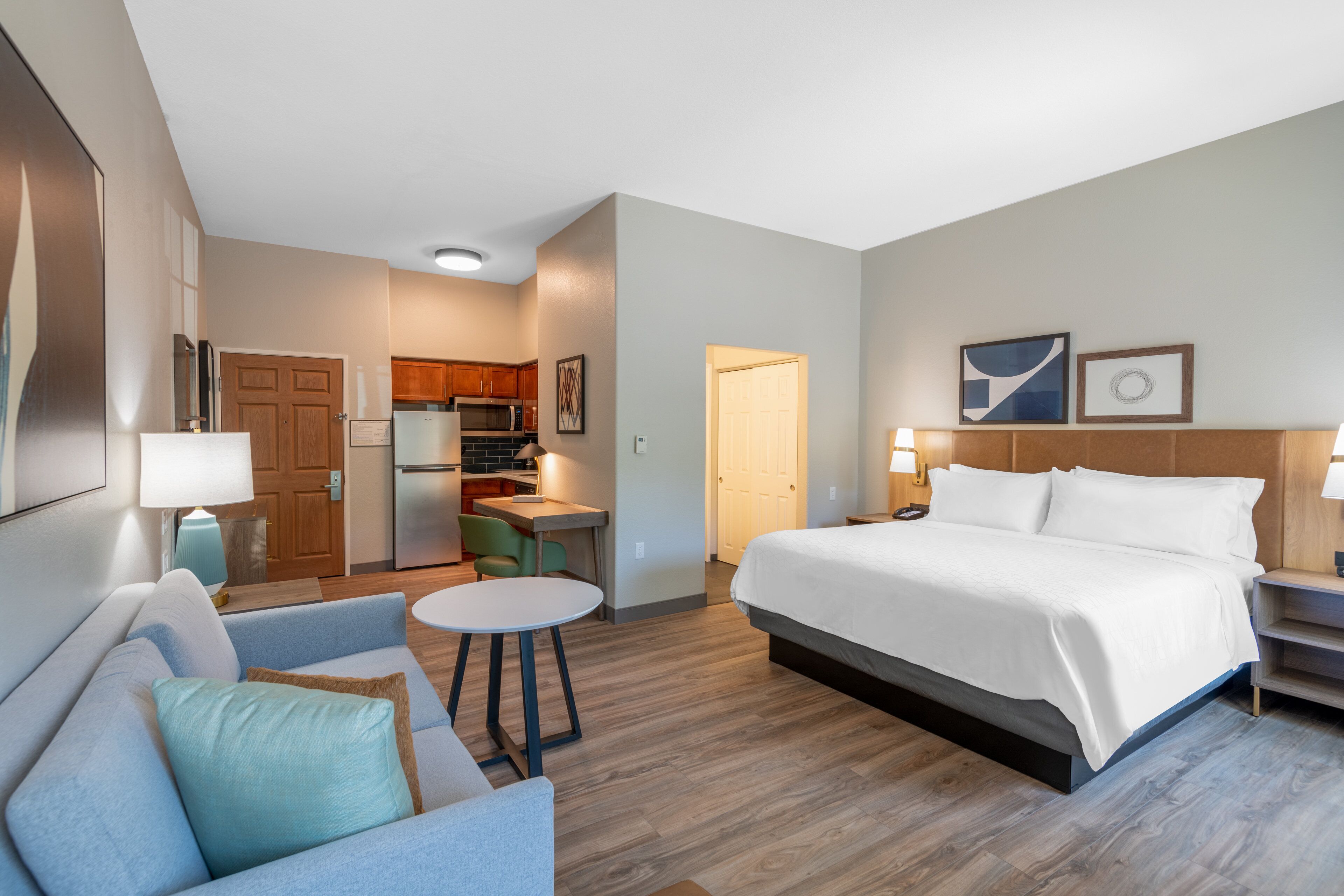 Quarto de hotel com cama, sofá, mesa pequena, escrivaninha e cozinha compacta.