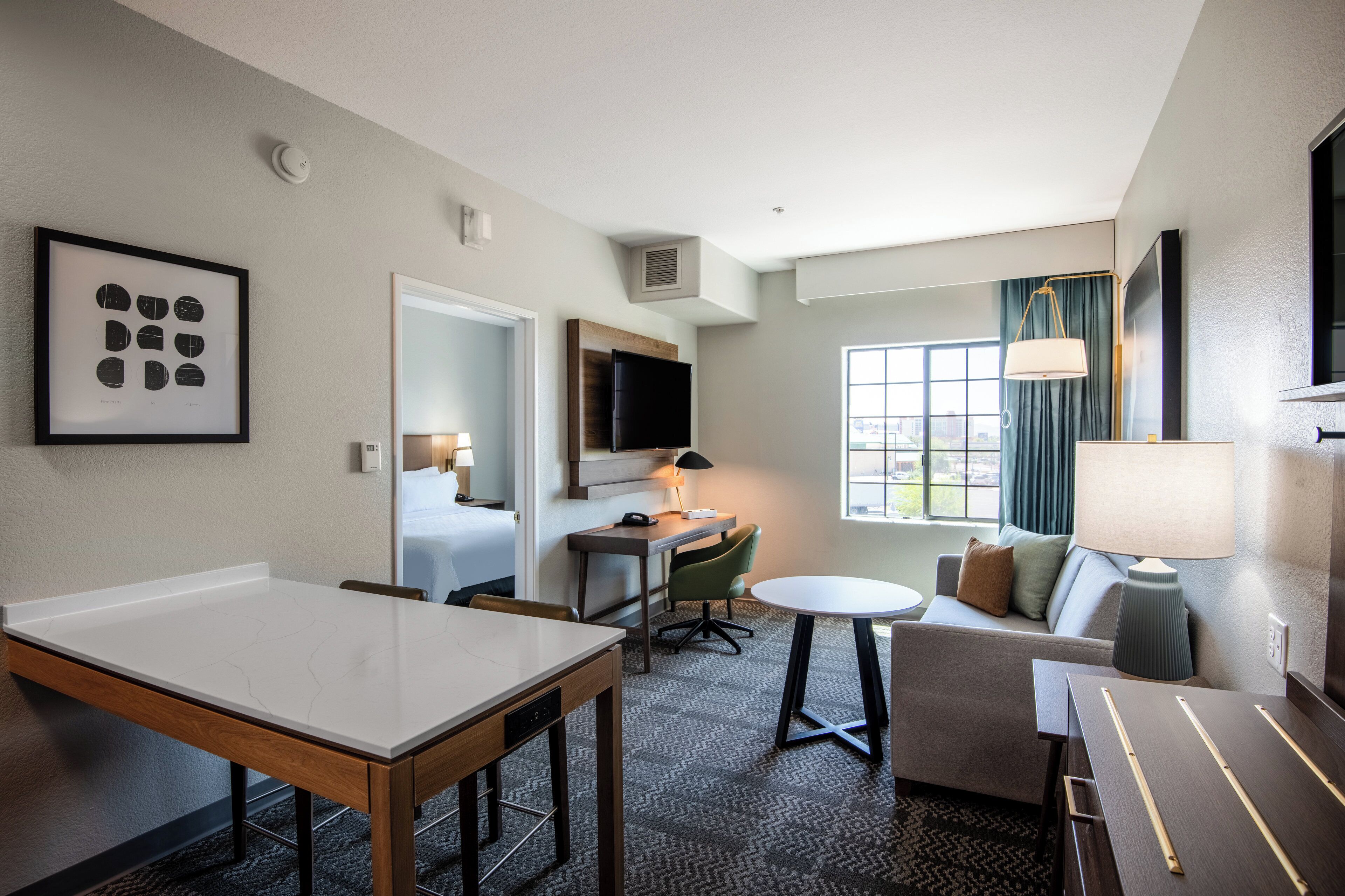A hotel room with a dining table, a sofa, a TV, and a window with curtains.