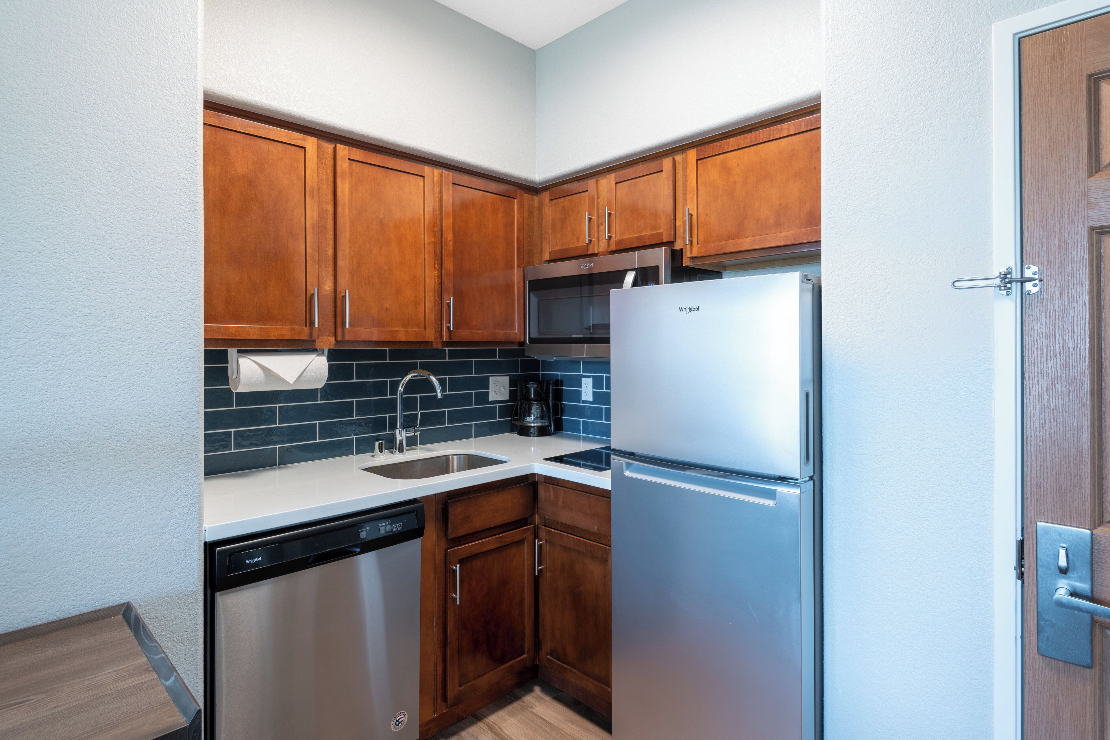 A compact kitchen with wooden cabinets, a stainless steel refrigerator, a dishwasher, and a coffee maker.