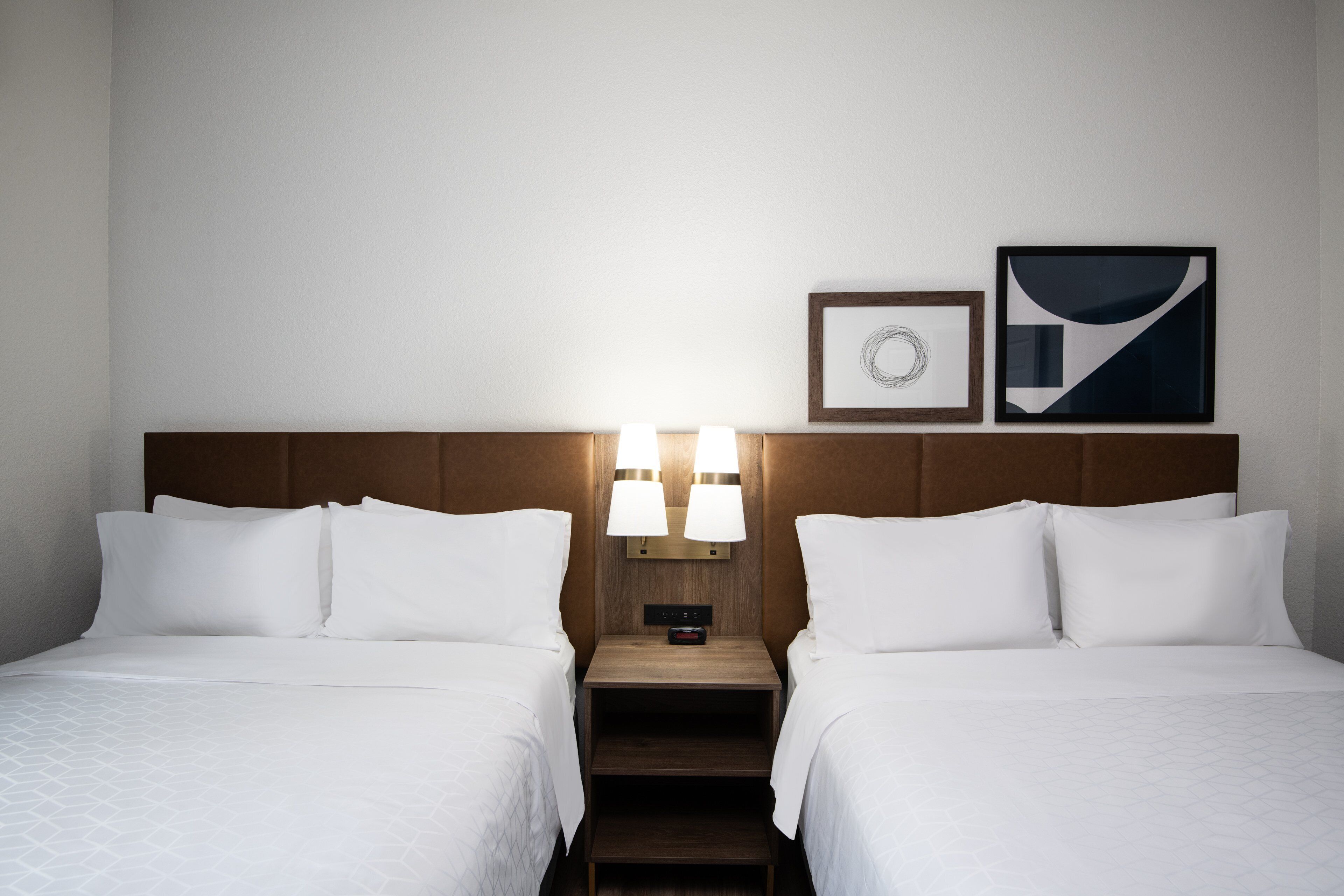 A hotel room with two beds, a wooden headboard, a nightstand with lamps, and framed artwork on the wall.