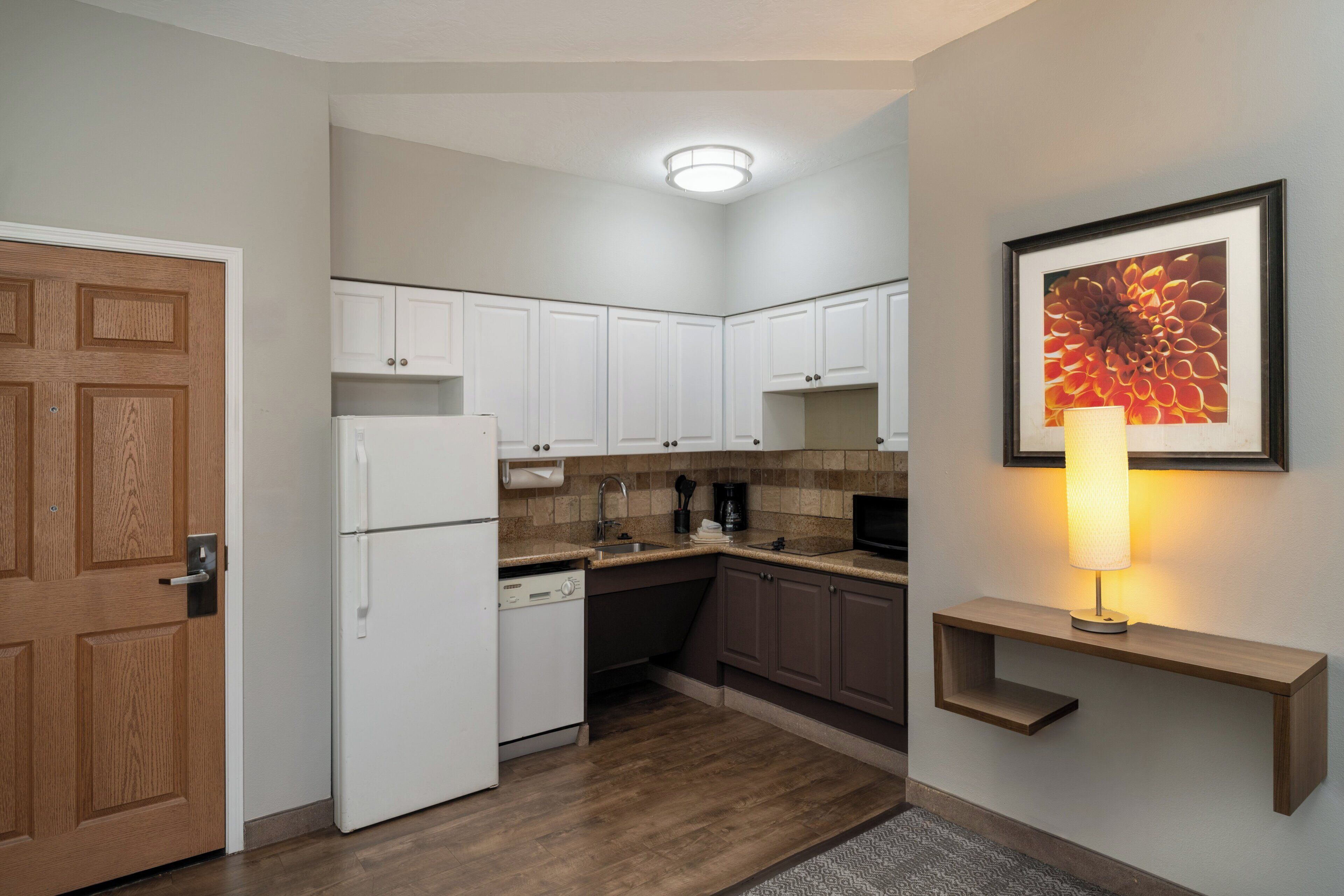 A compact kitchen with white cabinets, a refrigerator, a coffee maker, and a wall-mounted shelf with a lamp.
