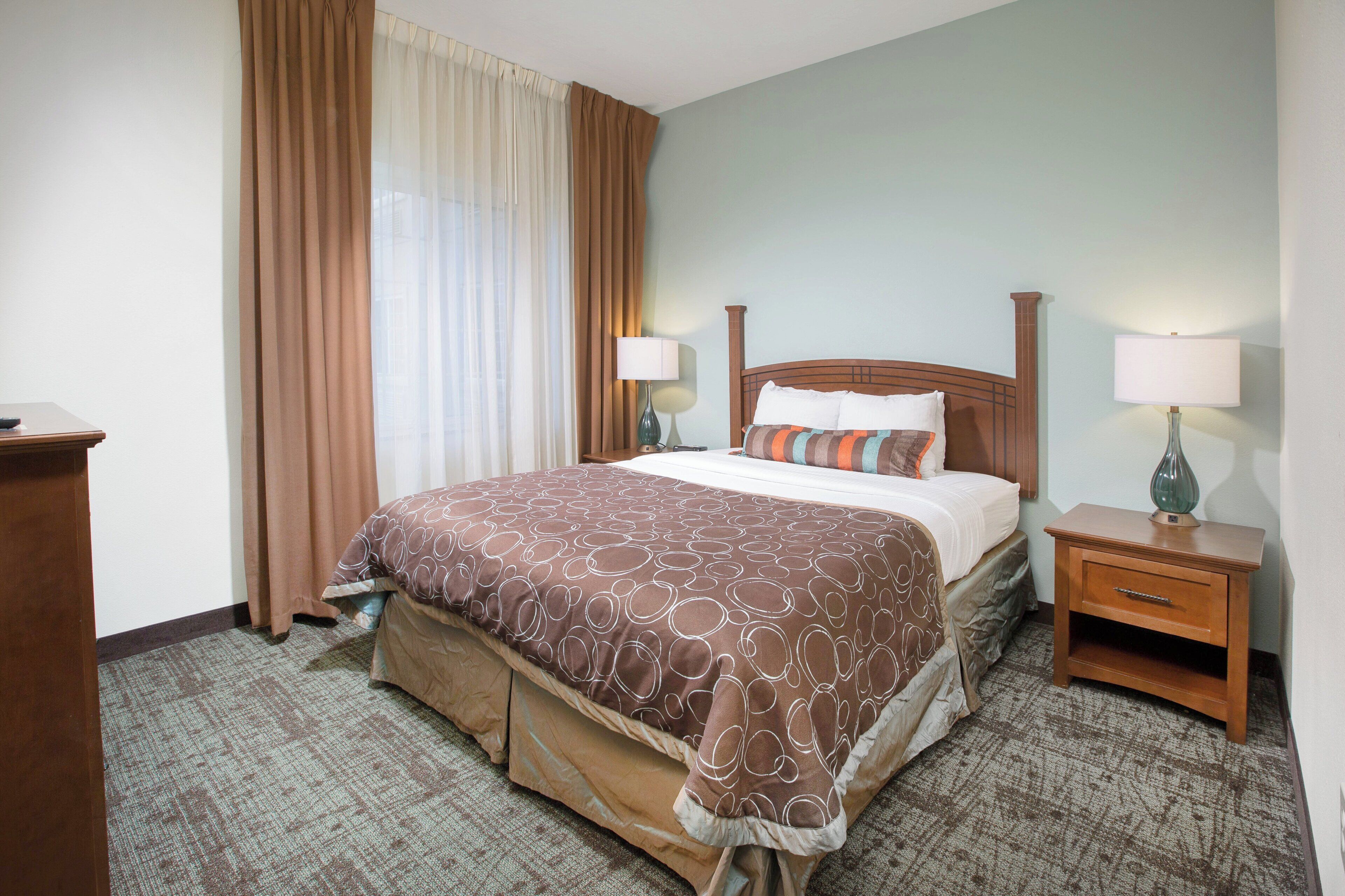 Une chambre d’hôtel comprenant un lit, deux tables de chevet, une commode, une fenêtre avec des rideaux et un sol recouvert de moquette.