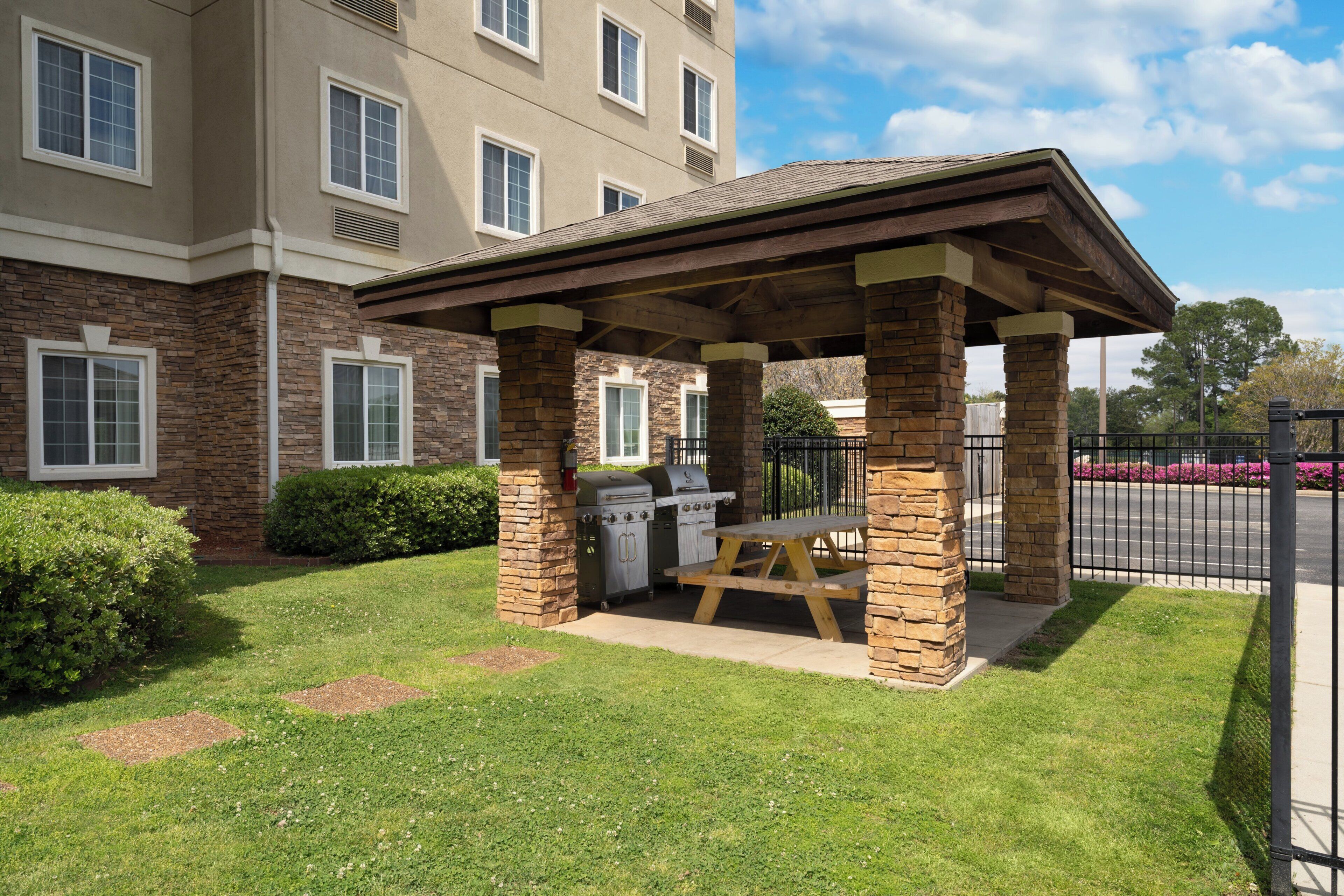 A covered outdoor grill area with stone pillars, a picnic table, and a brick building in the background.