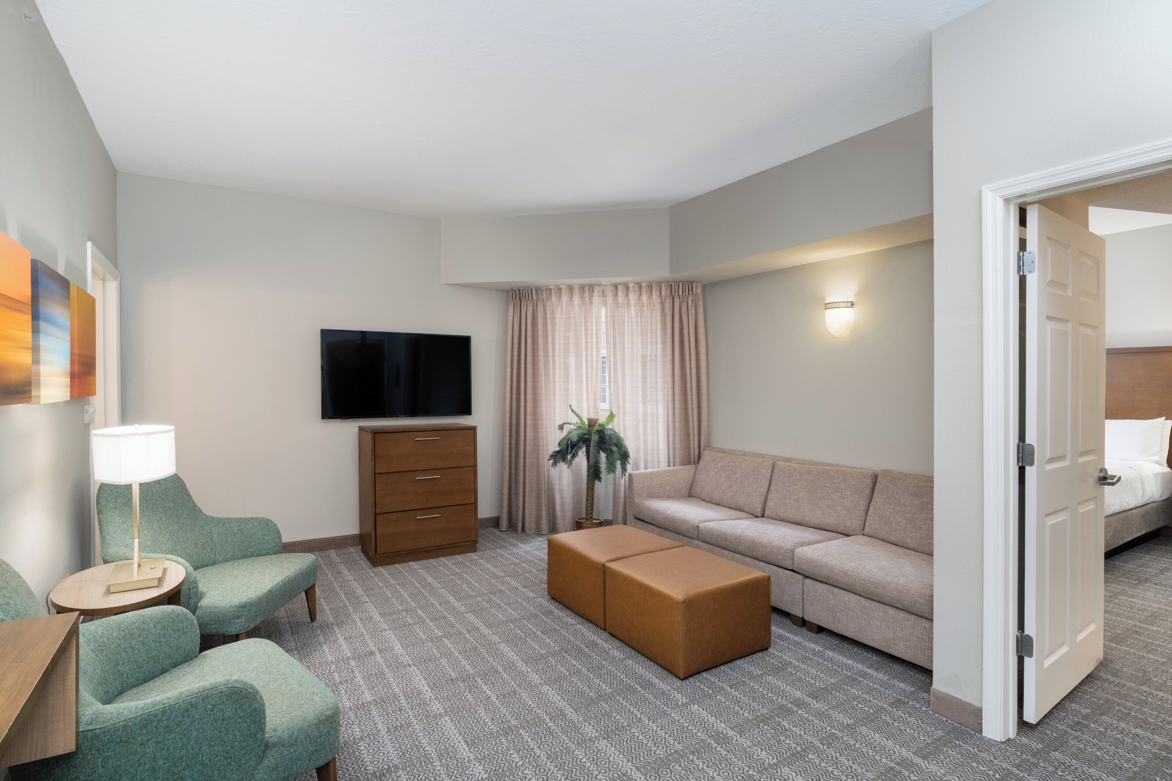 Une chambre d’hôtel comprenant un canapé, deux fauteuils, une télévision, une lampe, une plante et une porte donnant sur une autre pièce.