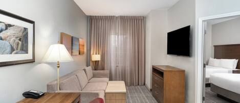 A hotel room with a sofa, a desk, a television, a bed, a lamp, and a window with curtains.