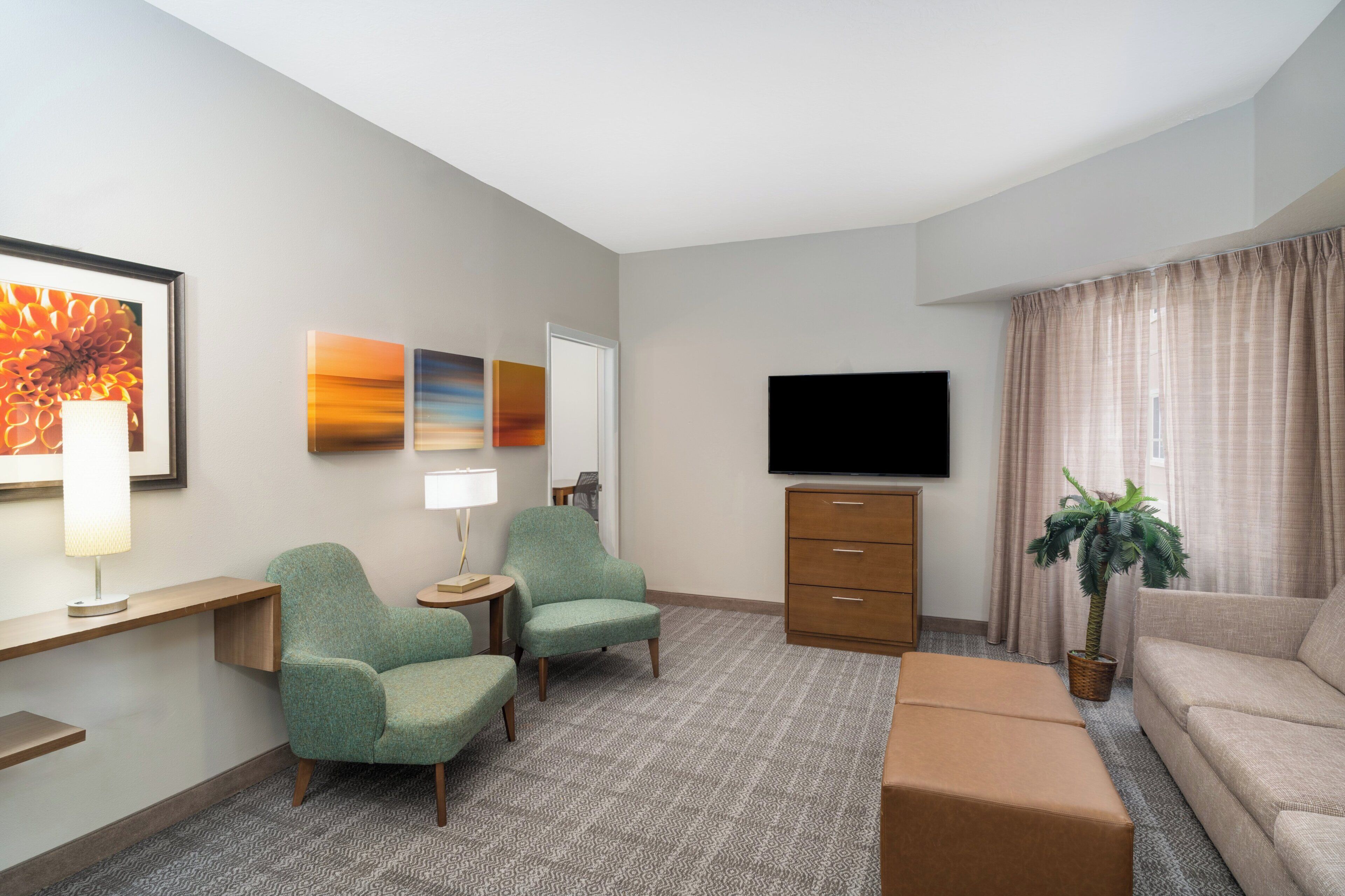 A hotel room with a flat-screen TV, two armchairs, a sofa, a small table with a lamp, a potted plant, and artwork on the walls.