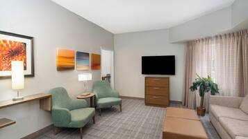 A hotel room with a flat-screen TV, two armchairs, a sofa, a small table with a lamp, a potted plant, and artwork on the walls.