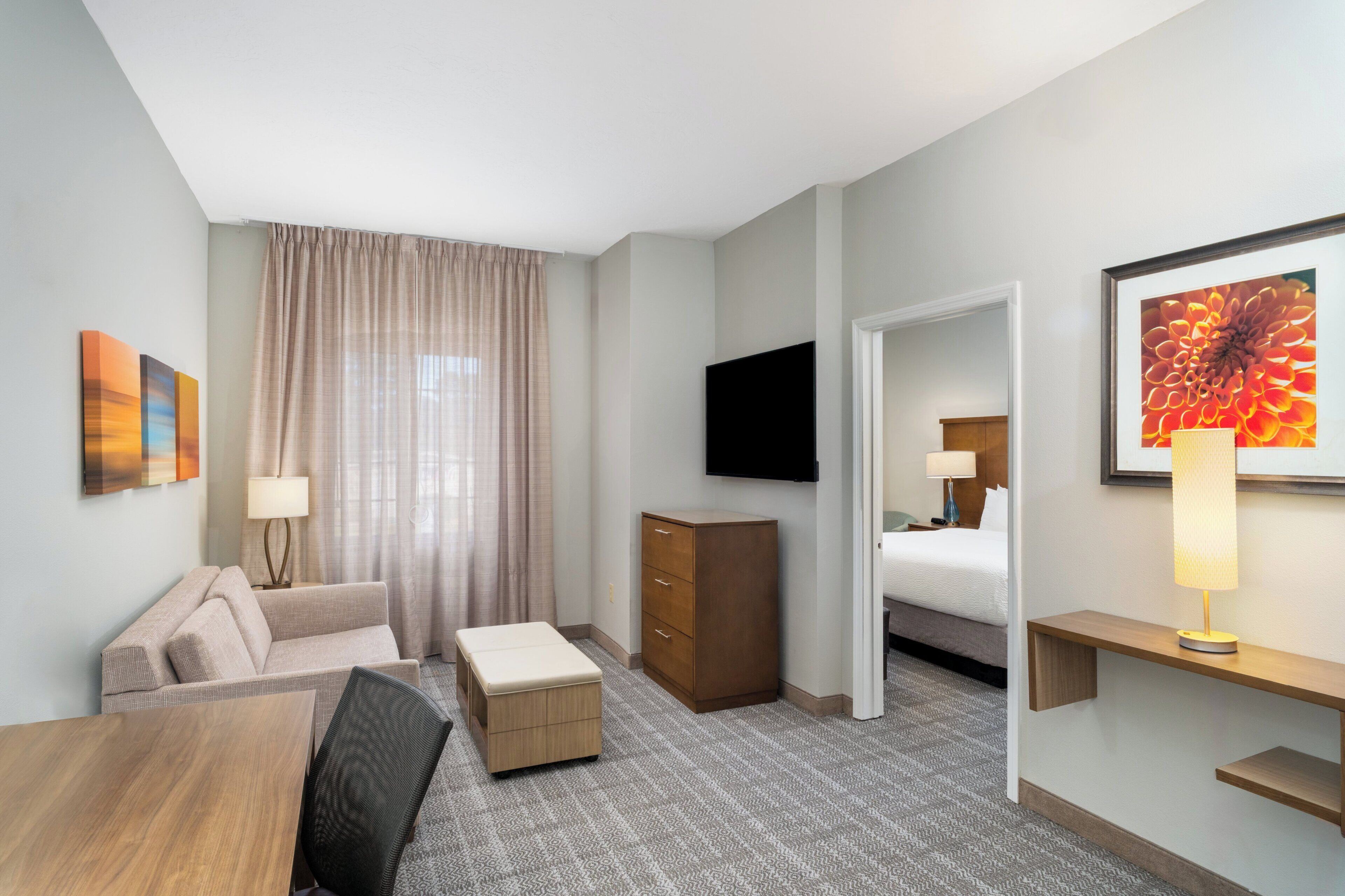 A hotel room with a sofa, a wooden desk, a television, a bed, a dresser, and a window with curtains.