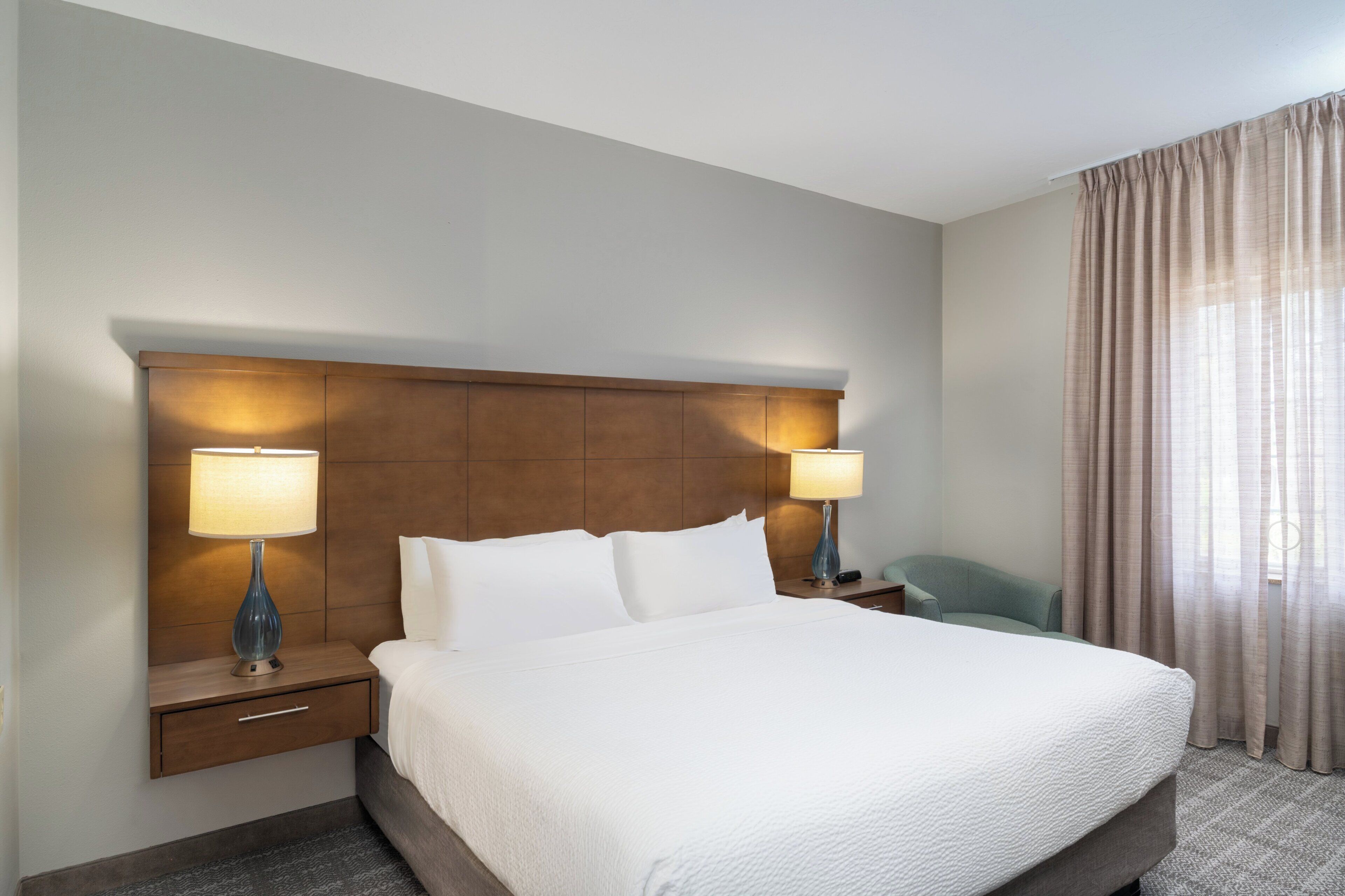 Une chambre d’hôtel avec un grand lit, deux lampes de chevet, une tête de lit en bois, une table de chevet avec un tiroir et une fenêtre avec des rideaux.