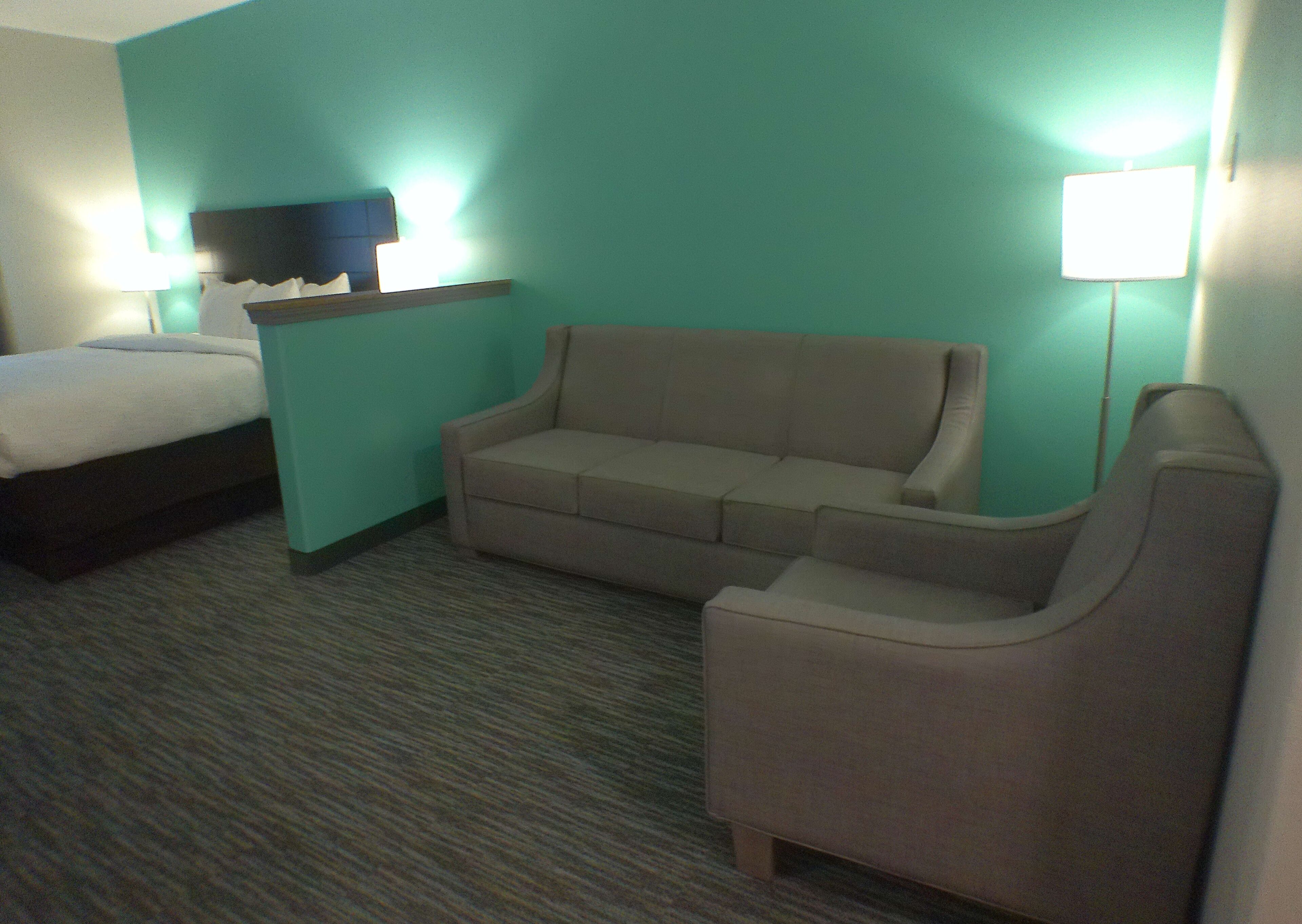 Habitación de hotel con cama, sofá, sillón, televisor sobre un soporte y lámpara.