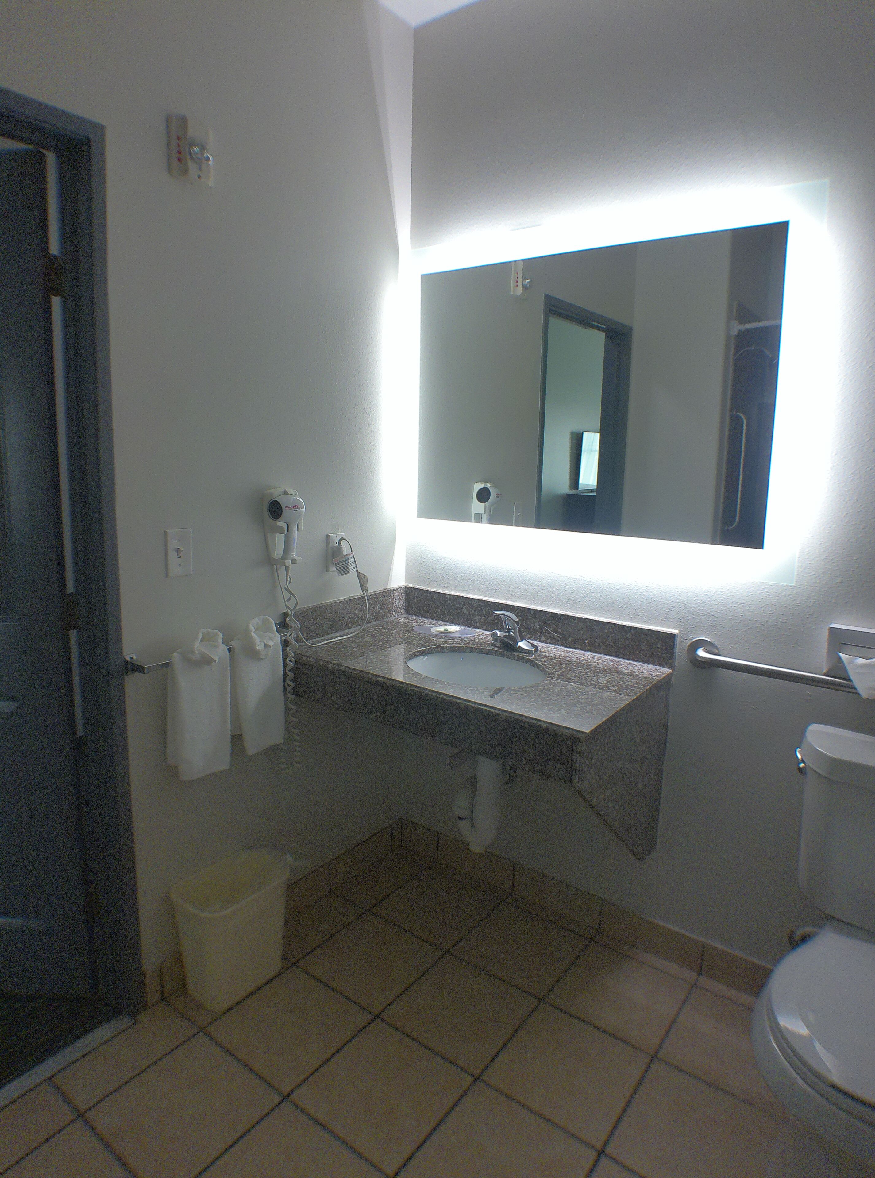 Ein Badezimmer mit Waschbecken, Spiegel und Toilette. Handtücher hängen an einem Wäscheständer, ein Mülleimer steht bereit und ein Handtrockner ist an der Wand angebracht.