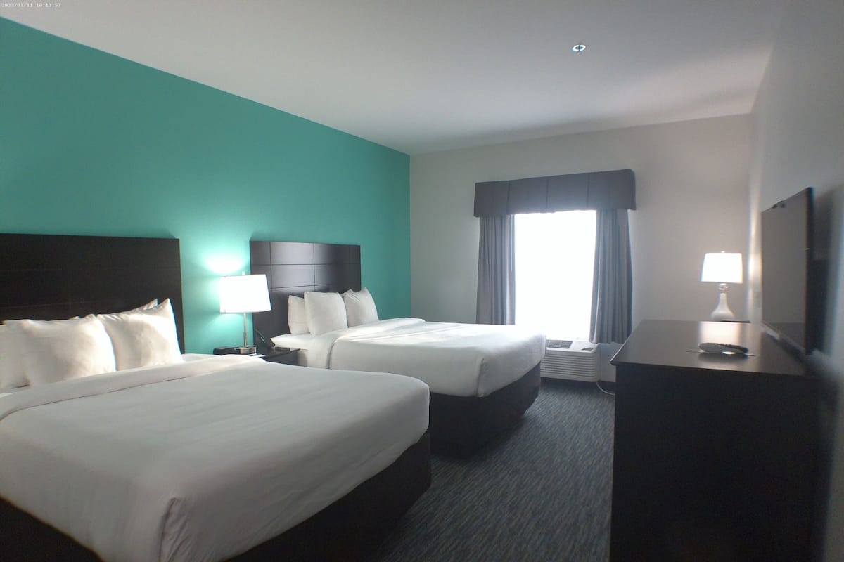 A hotel room with two beds, a desk, a lamp, and a large window with curtains.