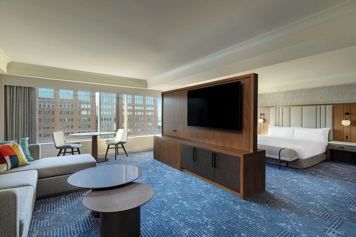 A hotel room with a large flat-screen TV, a bed, a sofa, a small table, and a view of the cityscape through large windows.