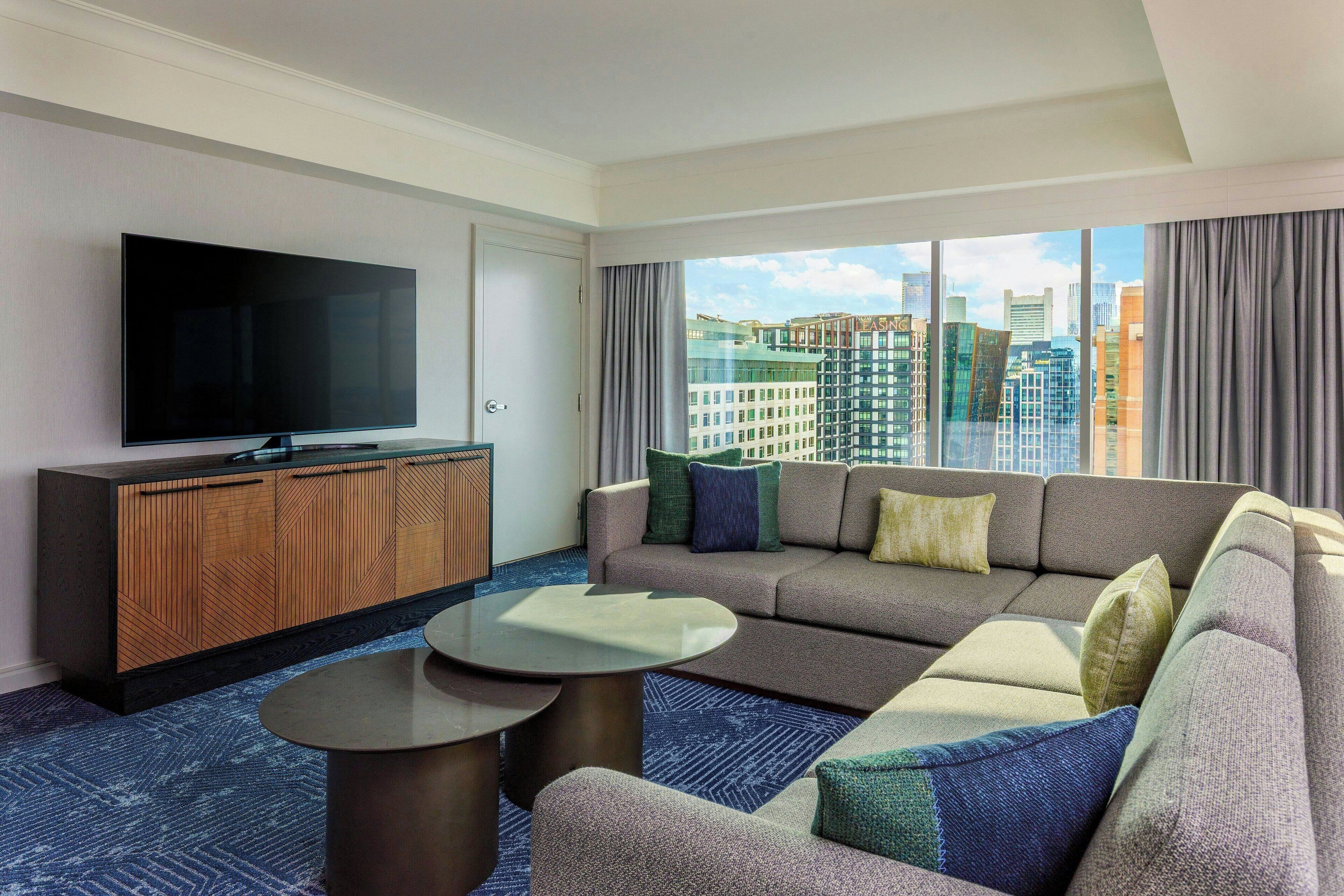 Habitación de hotel moderna con una amplia televisión de pantalla plana, un conjunto de sofás, una mesa de centro y vistas de la ciudad a través de grandes ventanales.