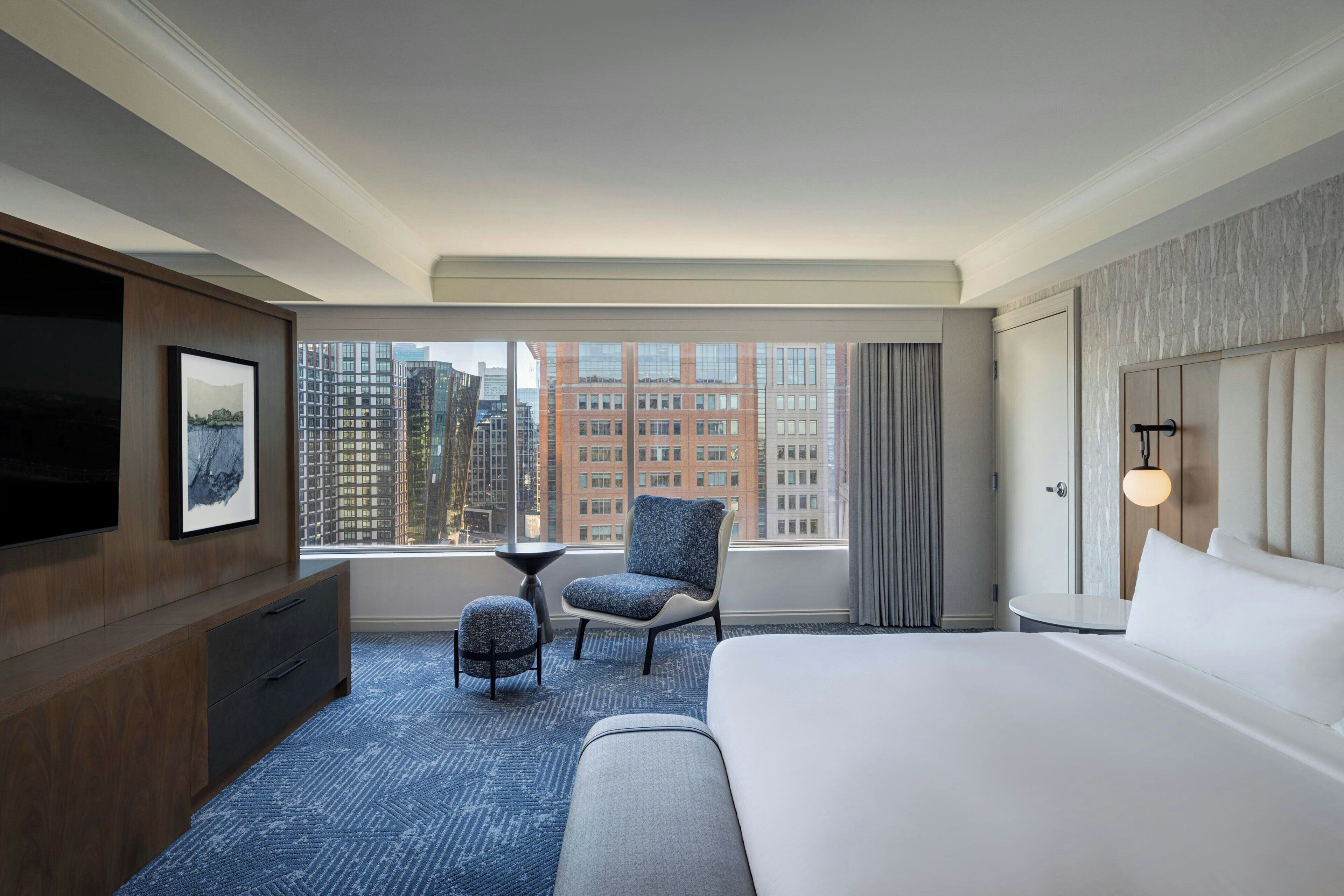 Habitación de hotel con una cama grande, un televisor montado en la pared, una mesita pequeña, una silla y vistas a la ciudad a través de la ventana.