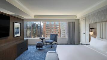 Habitación de hotel con una cama grande, un televisor montado en la pared, una mesita pequeña, una silla y vistas a la ciudad a través de la ventana.