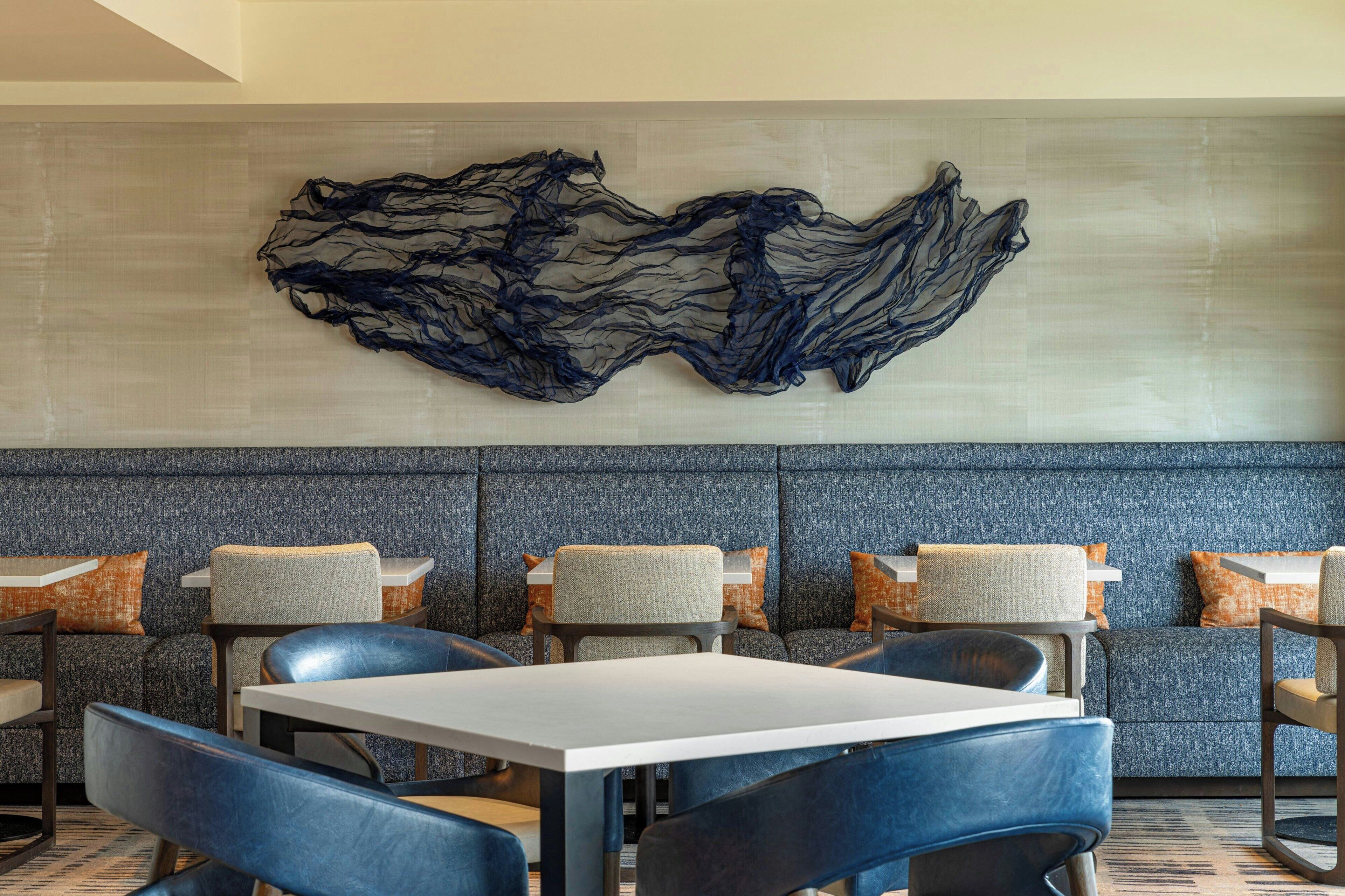 Un restaurante moderno con una zona de estar azul grisácea, mesas blancas y una gran obra de arte abstracta en la pared.