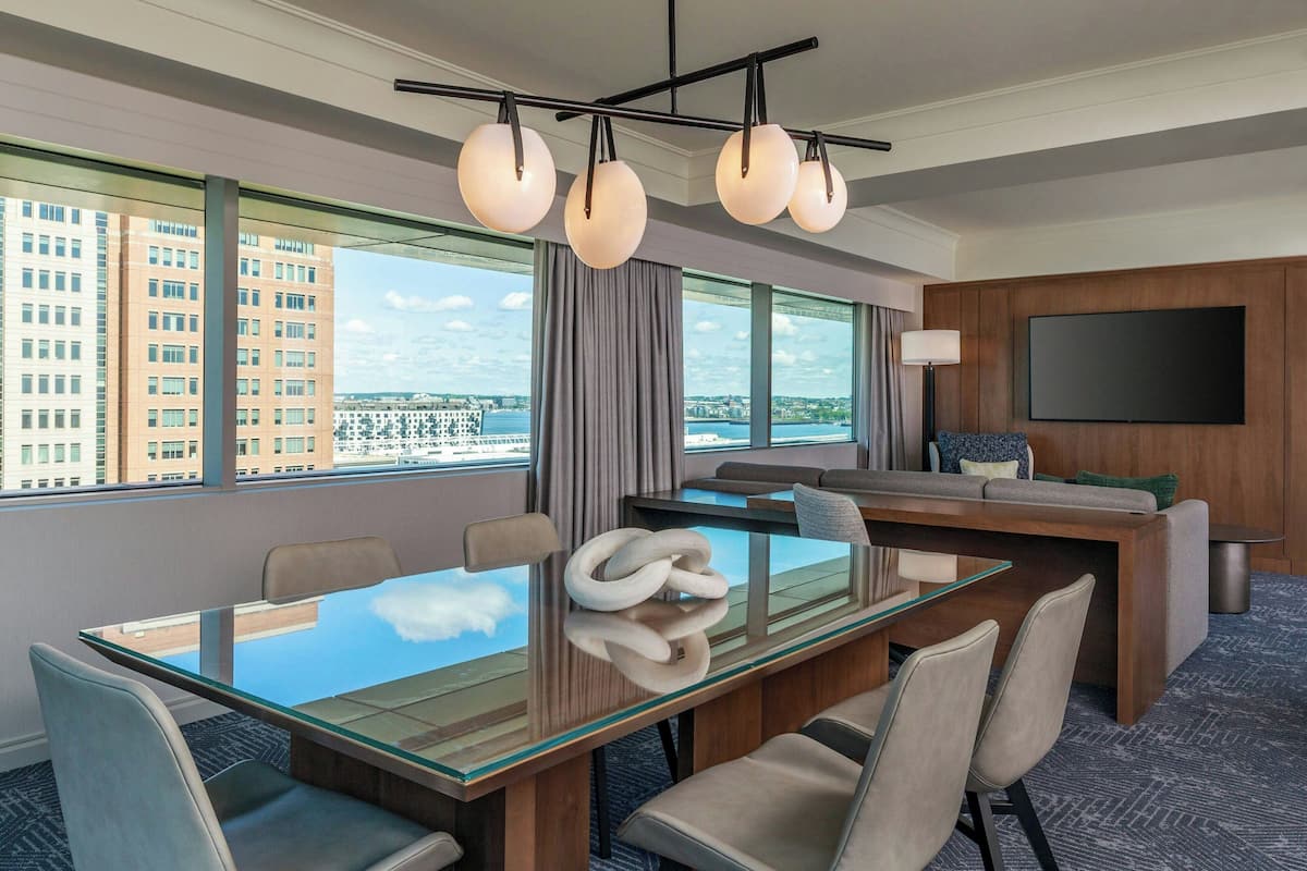 A modern hotel room with a large glass table, comfortable seating, a flat-screen TV, and large windows offering a city view.
