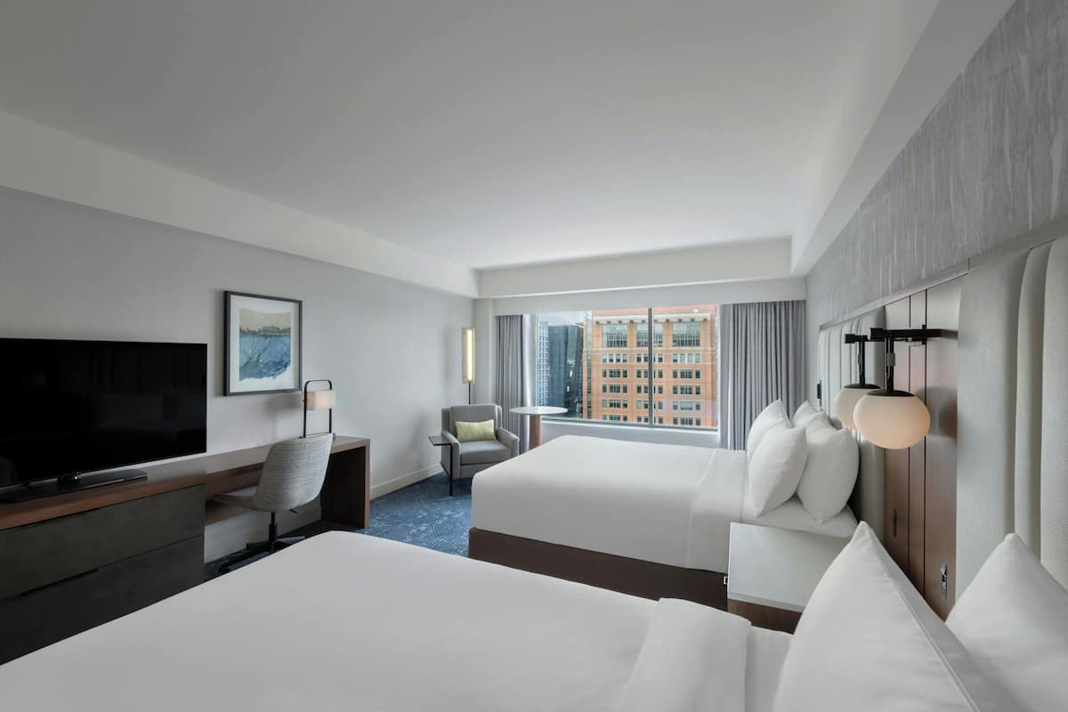 A hotel room with two beds, a desk with a TV, a chair, a window with a city view, and a wall-mounted light fixture.