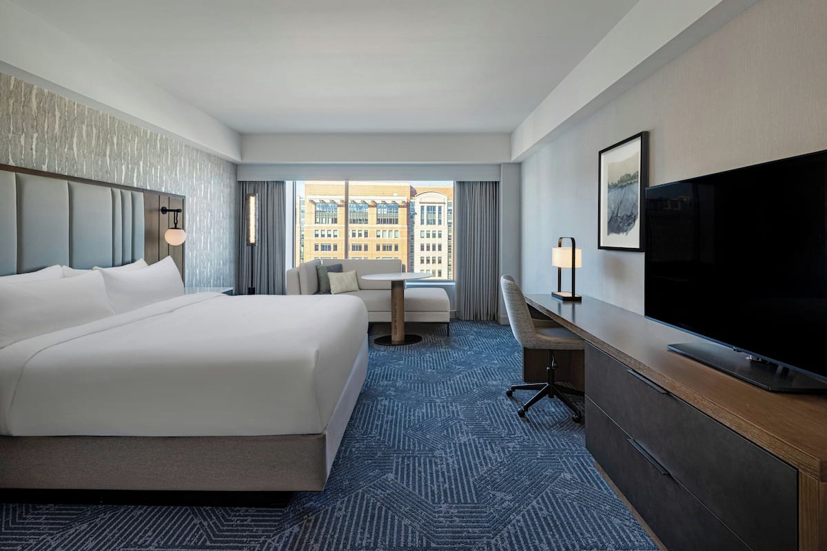 A hotel room with a large bed, a desk, a flat-screen TV, and a view of buildings through the window.