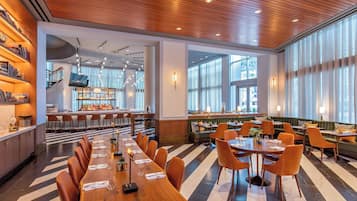 Un restaurante moderno con una larga mesa de madera, sillas anaranjadas y una zona de bar al fondo.
