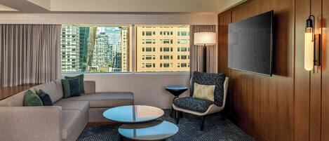 A modern hotel room with a large window, a sofa, two round tables, a chair, and a flat-screen TV mounted on the wall.