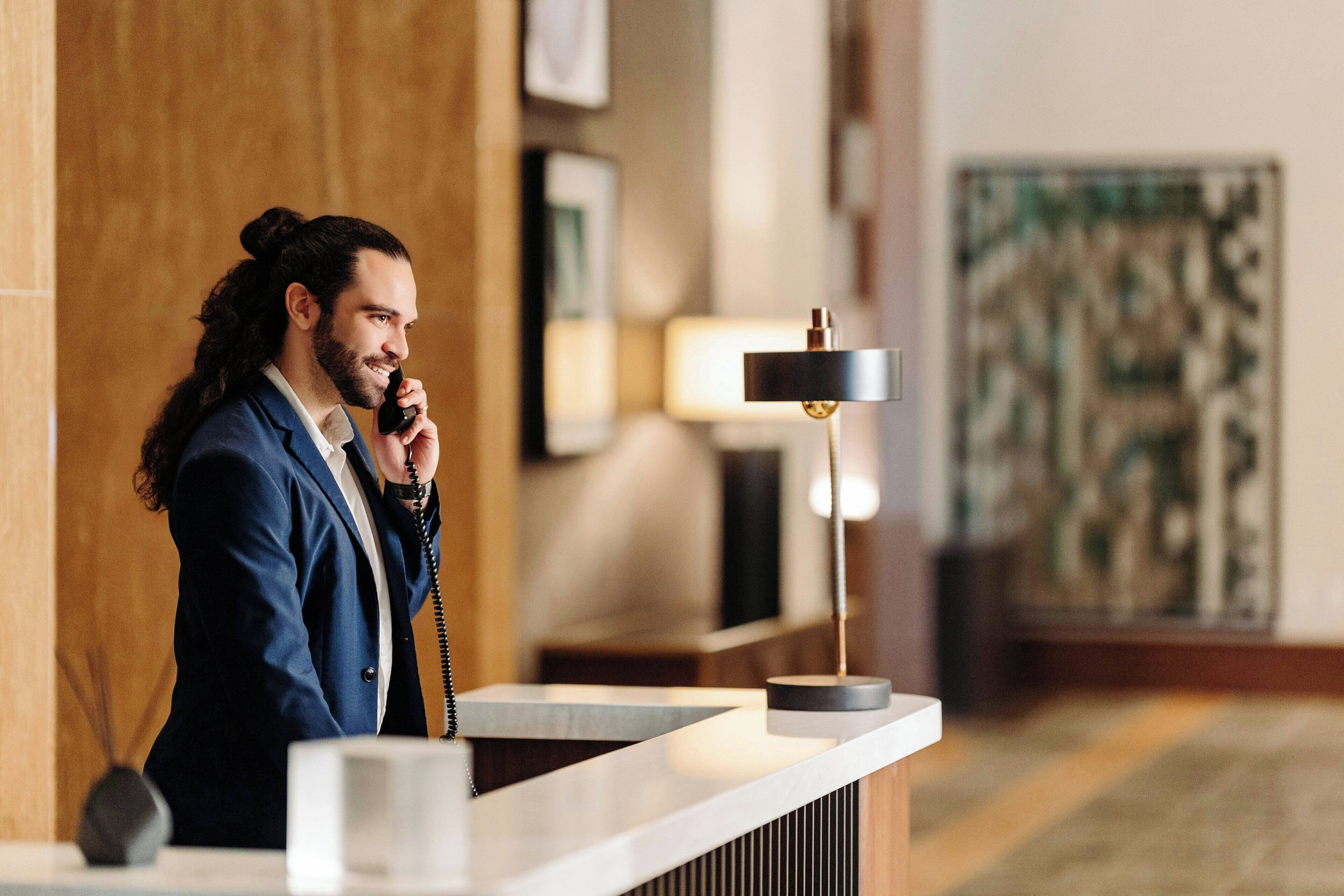 Un homme en costume se tient au comptoir de la réception d’un hôtel, avec un téléphone, une lampe et des œuvres d’art encadrées accrochées au mur.
