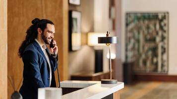 A man in a suit at a hotel reception desk with a telephone, a lamp, and framed artwork on the wall.