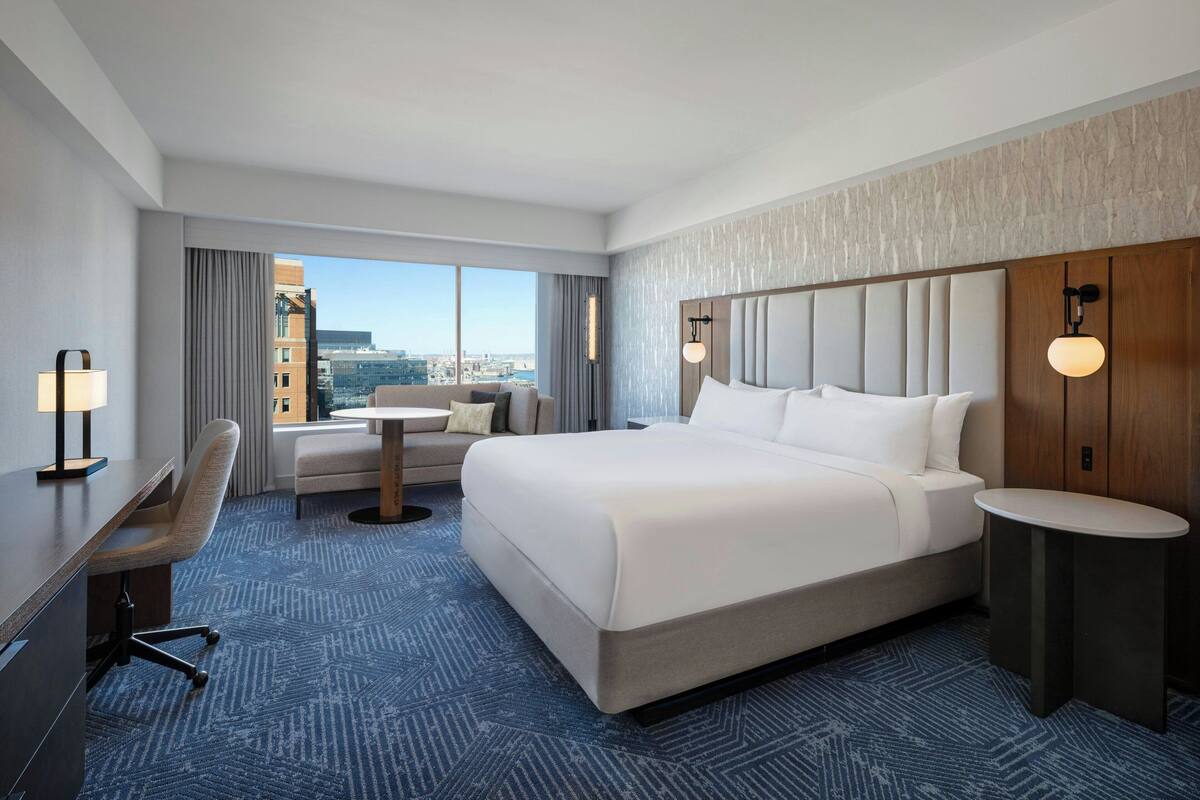 A hotel room with a large bed, a desk, a chair, a small table, and a view of the city through large windows.