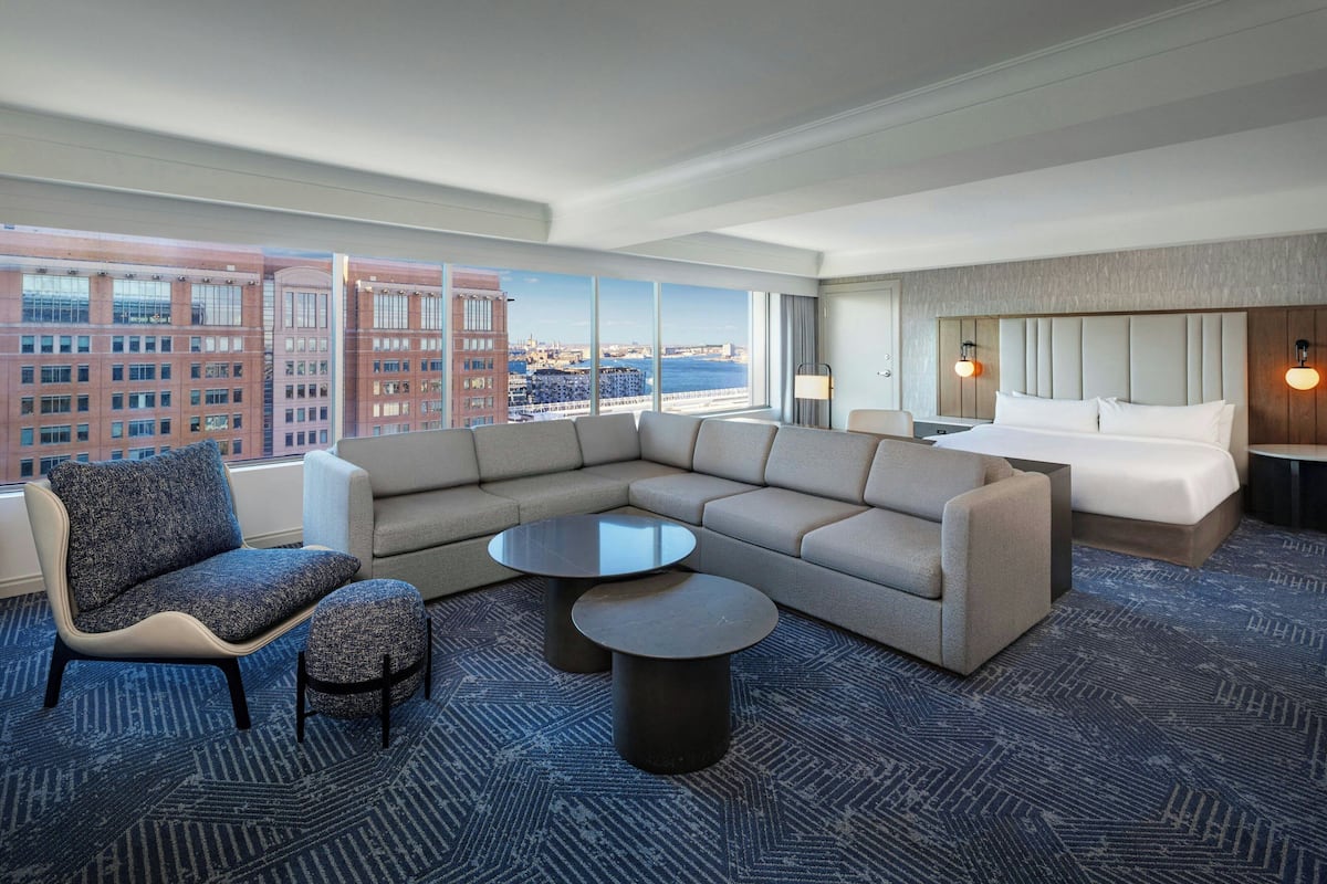 A modern hotel room with a large window offering a city view, a sofa, a chair, a small table, and a bed.