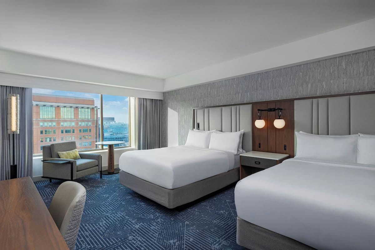 A hotel room with two beds, a desk, a chair, and a view of a cityscape.