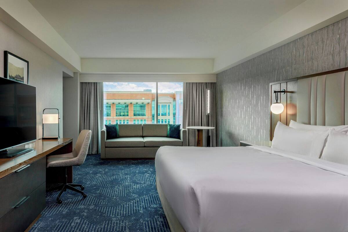 A hotel room with a large bed, a desk with a TV, a sofa, and a view of a building through the window.