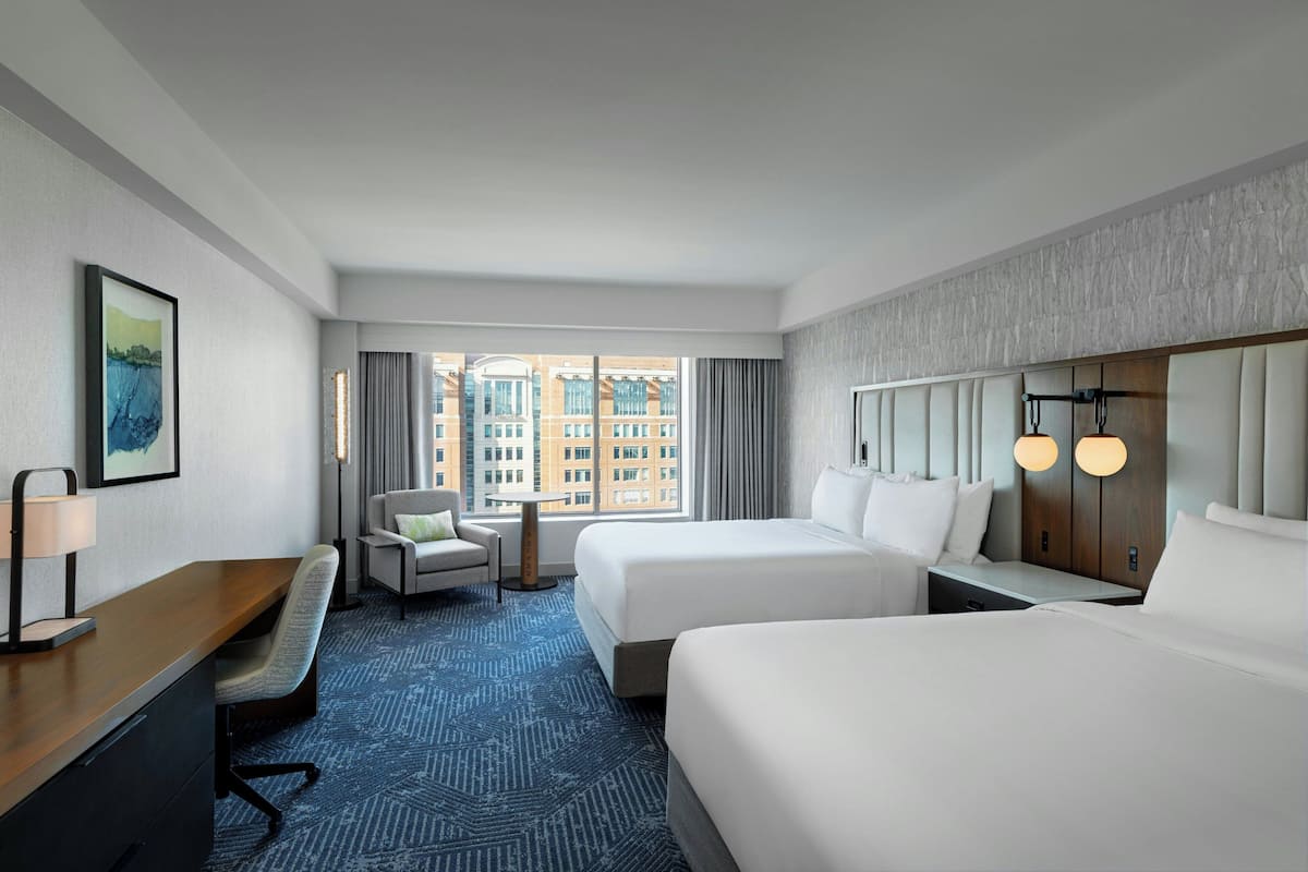 A hotel room with two beds, a desk, a chair, a small table, and a view of the city through large windows.