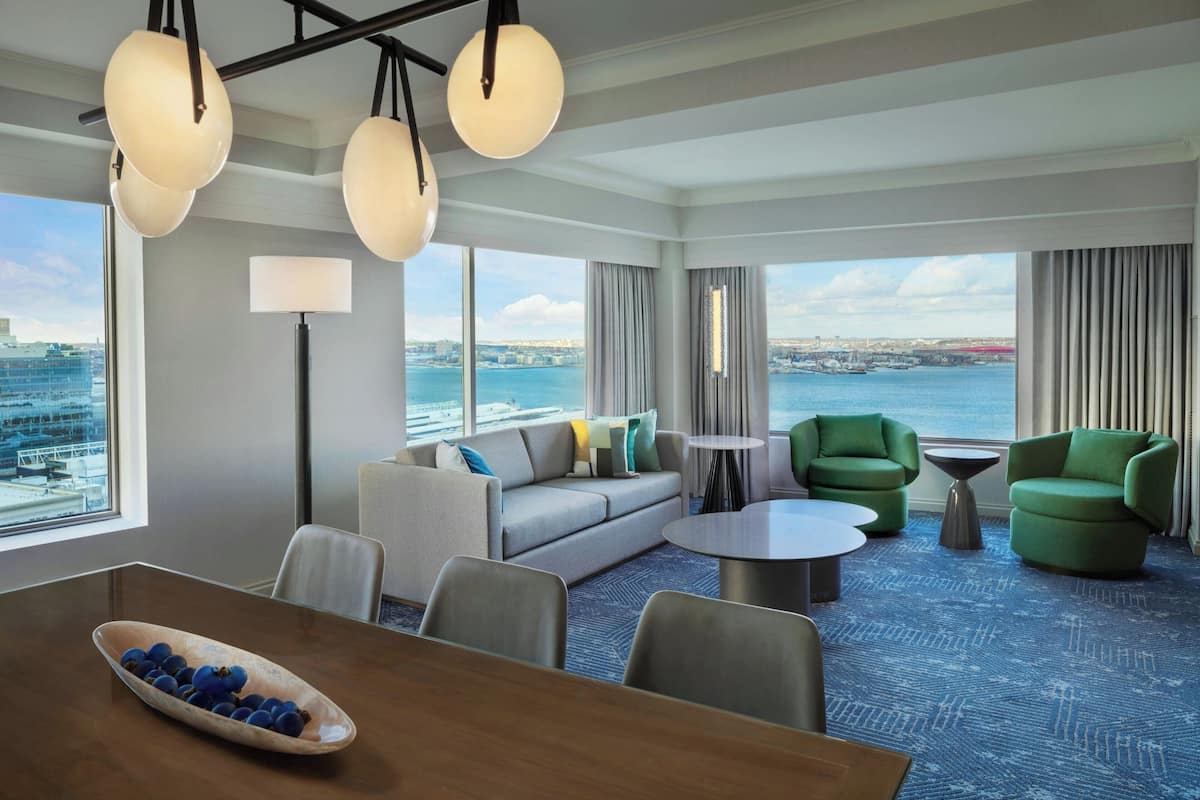 A modern hotel room with a large window offering a city view, a sofa set, green armchairs, a dining table with a bowl of fruit, and a patterned carpet.