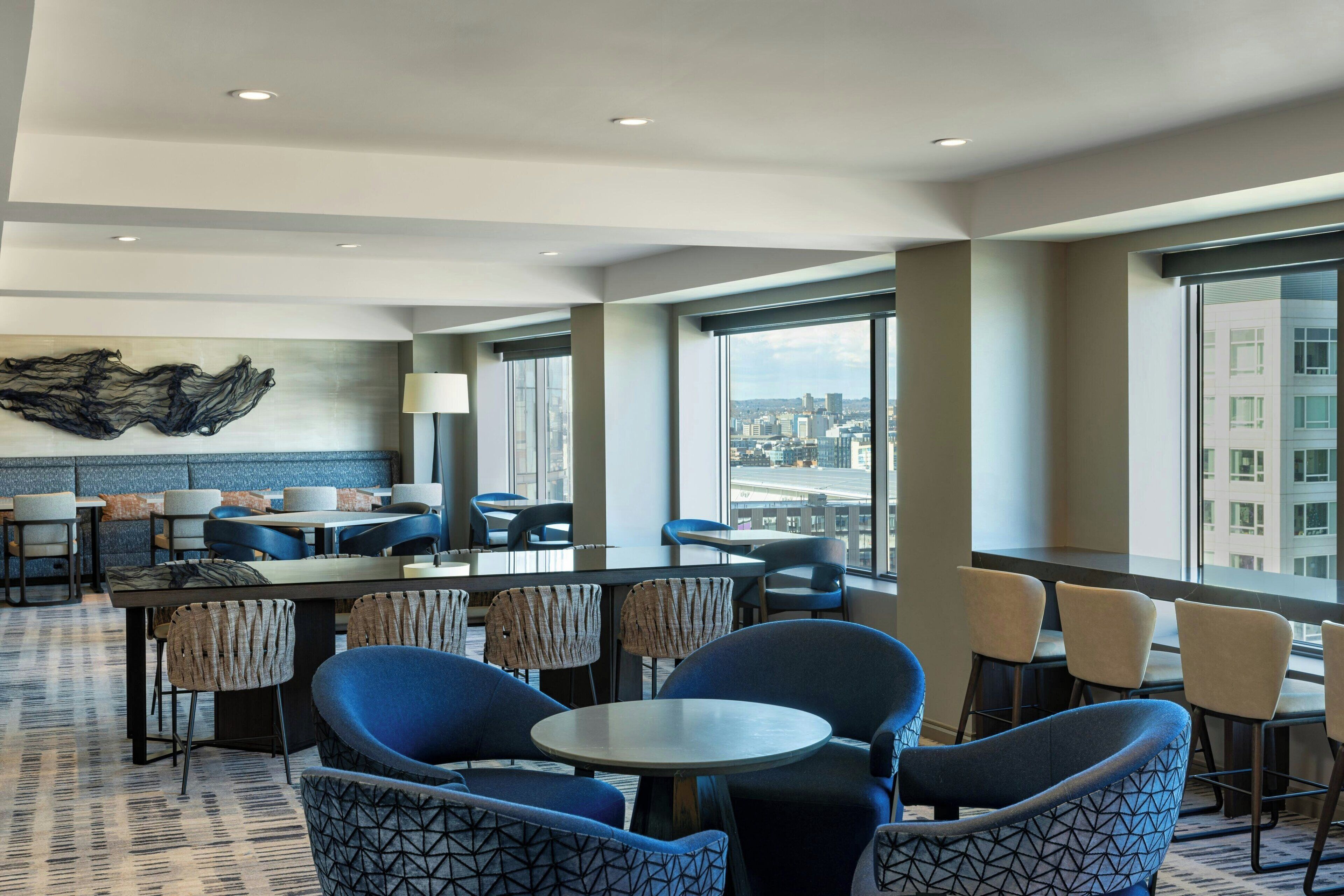 Een moderne hotellobby met een hoge plafond, grote ramen met uitzicht op de stad, en een gevarieerde zithoek bestaande uit tafels en stoelen.