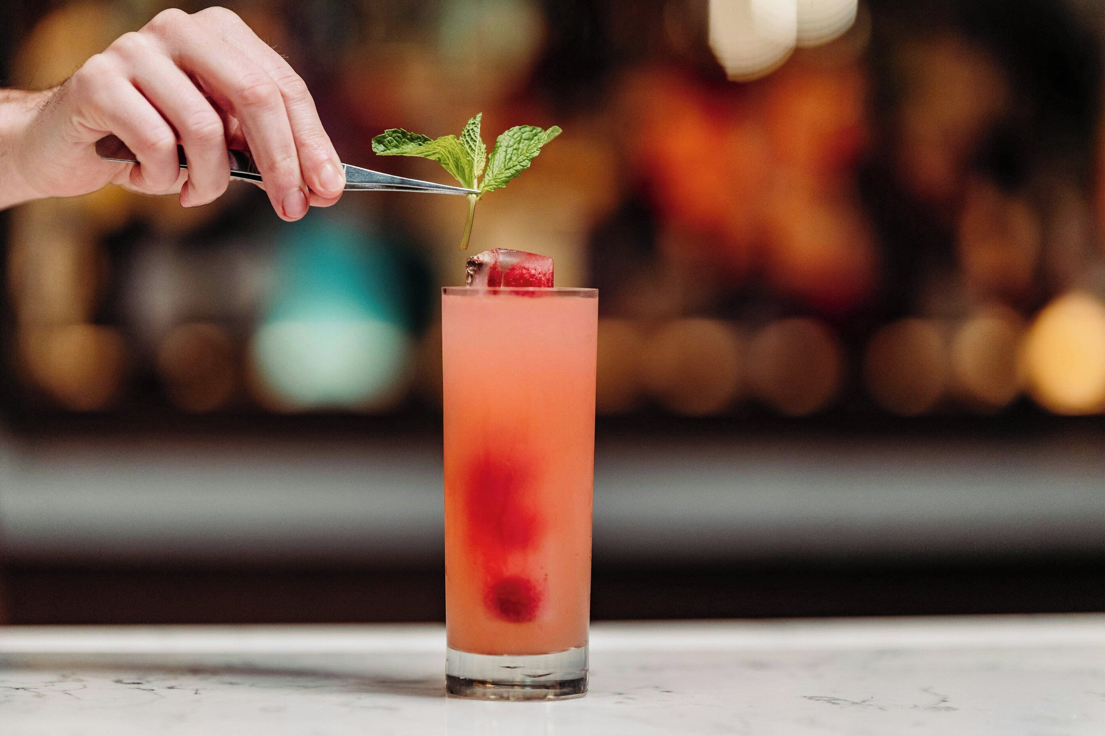En hånd placerer et mynteblad på en cocktail med et kirsebær.