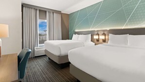 Habitación de hotel con dos camas, un escritorio, una silla, un ventanal con cortinas y una pared con diseño geométrico.