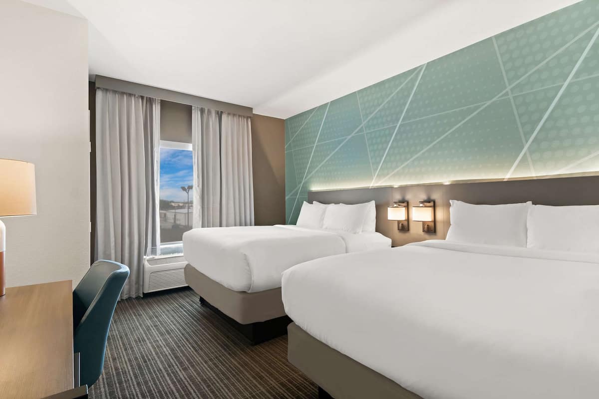 A hotel room with two beds, a desk, a chair, a window with curtains, and a geometric-patterned wall.