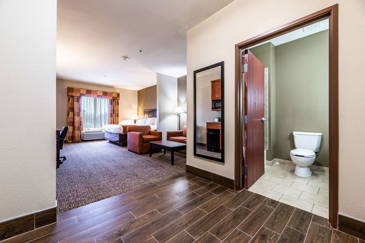A hotel room with a bed, two armchairs, a desk, a mirror, and a bathroom with a toilet.