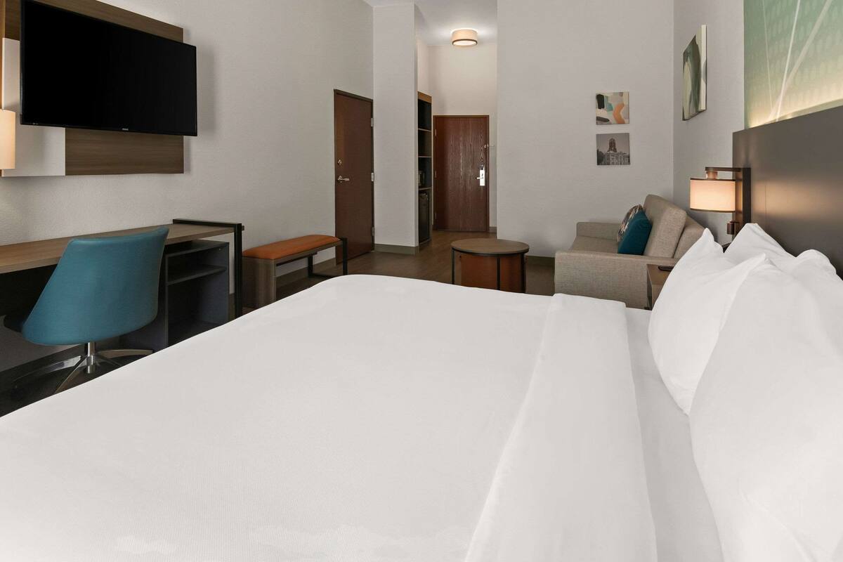 A hotel room with a bed, a desk with a chair, a small round table, a sofa, and a TV mounted on the wall.