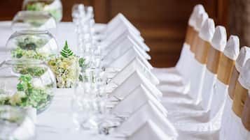 A long table with white tablecloths, glassware, and white napkin holders with gold bands.