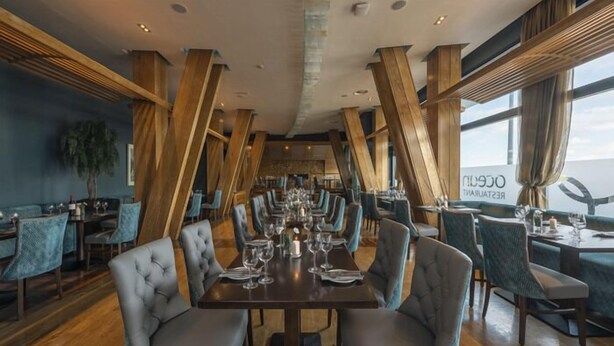 A restaurant with a modern interior, featuring high wooden beams, grey upholstered chairs, and a long dining table set for service.