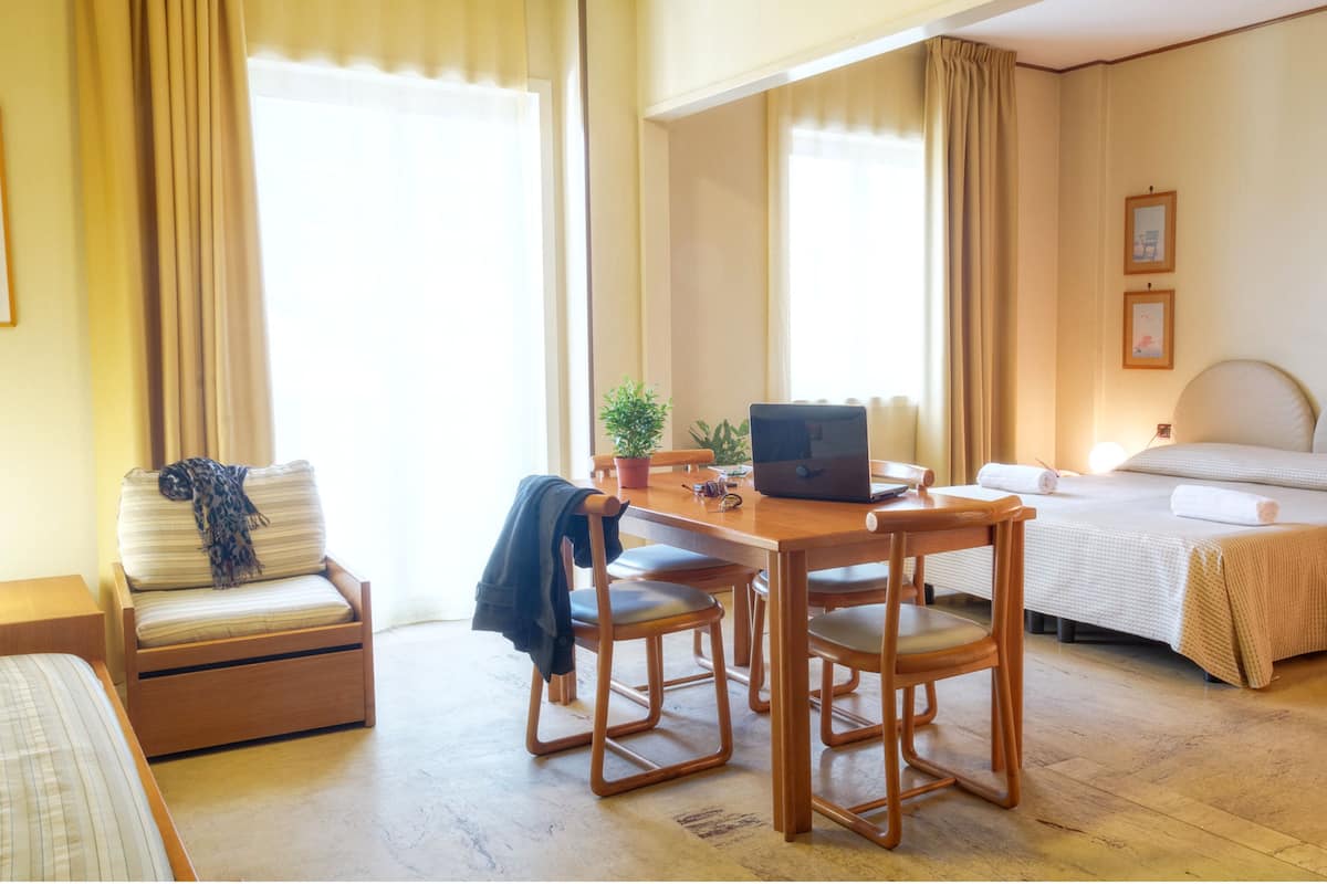 A hotel room with a bed, a desk with a laptop, a chair, a small table, a potted plant, and a window with curtains.
