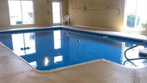 An indoor swimming pool with clear water, a white chair, and windows allowing natural light. - Randolph Inn and Suites (Winchester)
