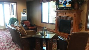 A living room with a fireplace, a television mounted above it, and a carpeted floor. - Randolph Inn and Suites (Winchester)