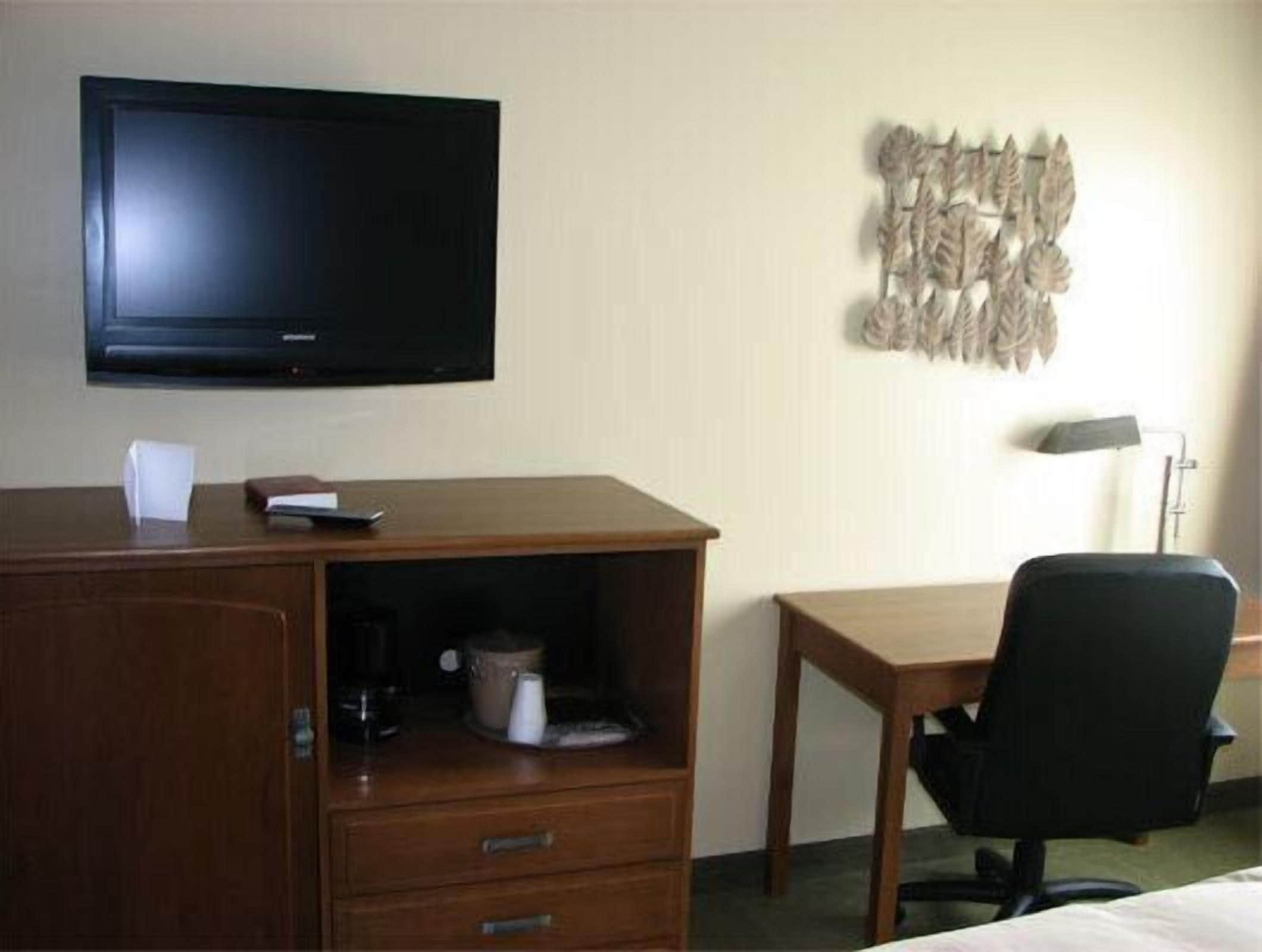 A hotel room with a wooden desk, a black office chair, a television mounted on the wall, and a decorative wall hanging.