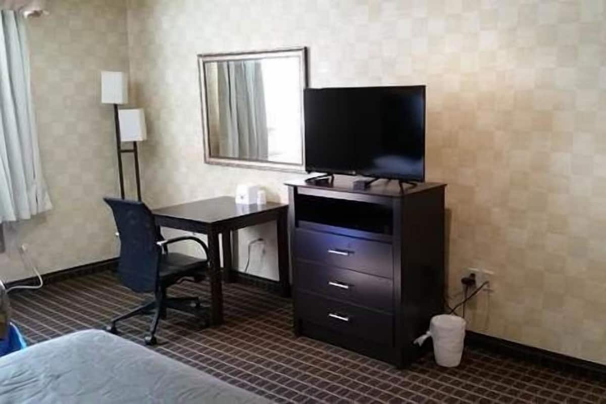 A hotel room with a bed, a desk with a chair, a television on a stand, a mirror, and a tiled floor.